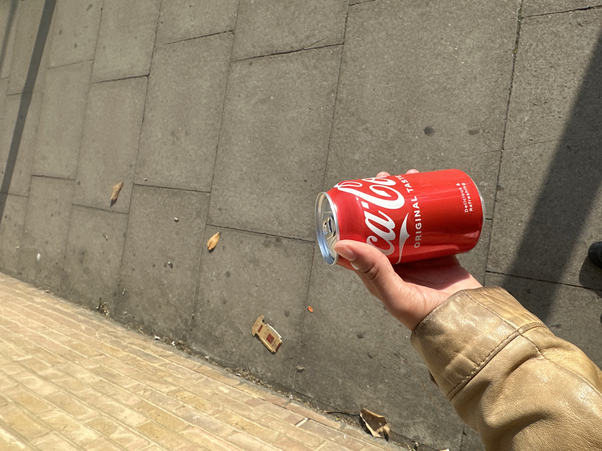 A hand in a tan leather jacket holds a red Coca-Cola can against a backdrop of grey paving stones and yellow brick. A piece of litter is on the grey paving stones. The location is possibly in London, United Kingdom.