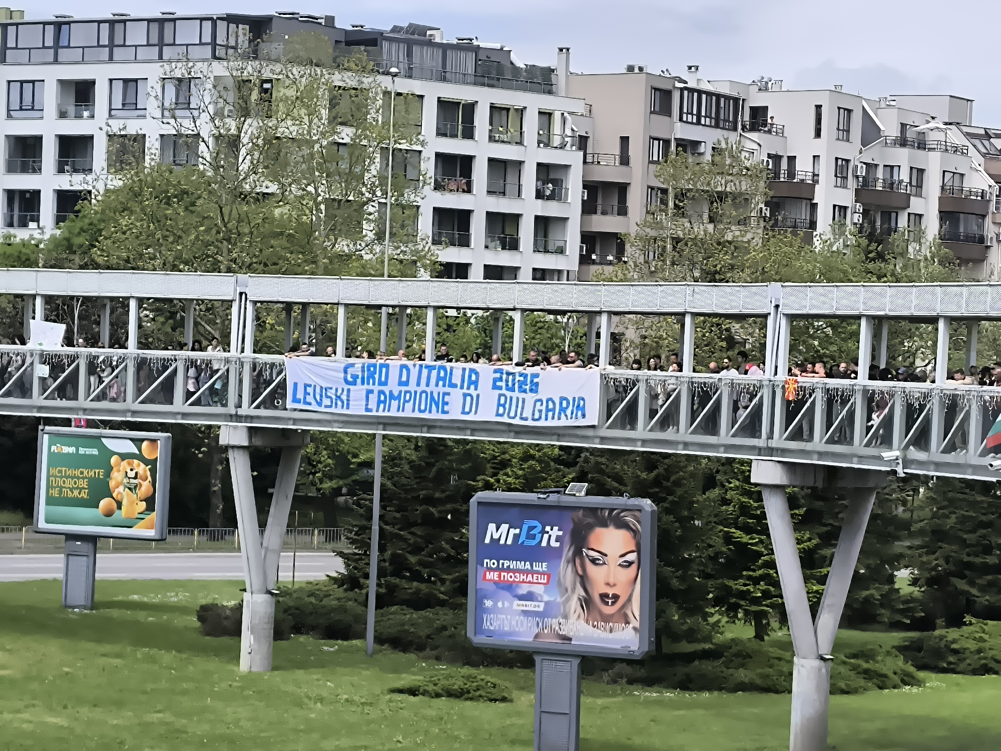The image shows a pedestrian bridge over a road in Burgas, Bulgaria. On the bridge, a large group of people are gathered, holding a banner that reads "GIRO D'ITALIA 2026 LEVSKI CAMPIONE DI BULGARIA." Below the bridge, on either side of the road, are billboards. One billboard advertises MrBit, a gambling site, featuring an image of a woman with heavy makeup and the tagline "ПО ГРИМА ЩЕ МЕ ПОЗНАЕШ" which translates to "You'll recognize me by my makeup." The other billboard promotes fruit juice, with the phrase "ИСТИНСКИТЕ ПЛОДОВЕ НЕ ЛЪЖАТ" which translates to "The real fruits don't lie." The background features multi-story residential buildings and trees. The day appears overcast.