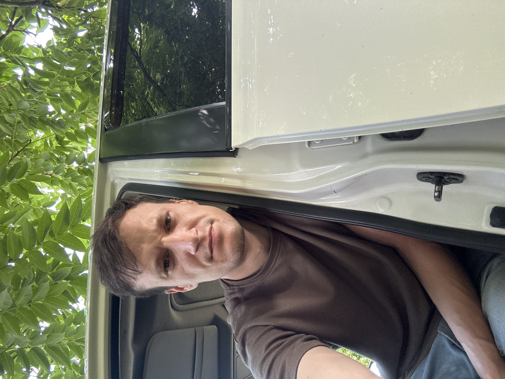 A man with short brown hair and a light complexion is sitting in a vehicle with the door open. He is wearing a brown t-shirt and blue jeans. The man is looking directly at the camera. The interior of the vehicle appears to be gray. The exterior of the vehicle is light-colored, possibly white or cream. Green foliage from a tree is visible above and behind the vehicle, suggesting an outdoor setting. The scene is set in Makiv, Ukraine.