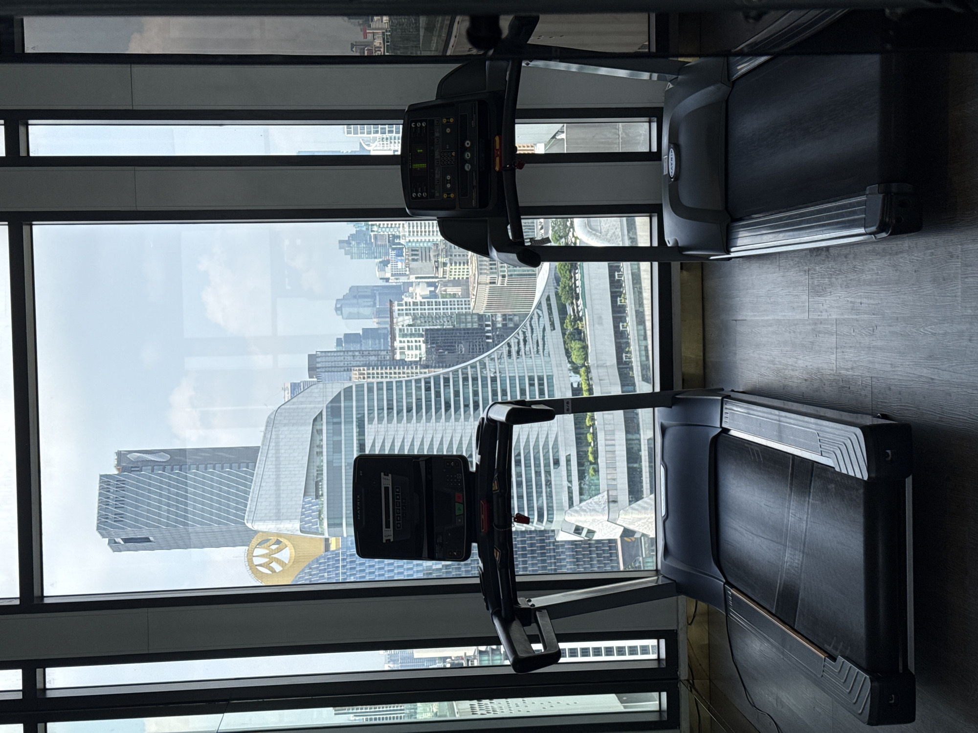 The image captures a scene inside a high-rise building, presumably a gym, with two treadmills positioned in front of a large window. The window offers a clear view of the Bangkok skyline, prominently featuring the distinctive architecture of the King Power MahaNakhon building with its pixelated design and the adjacent curved structure of the Central Embassy mall. The day appears bright with scattered clouds. The treadmills are dark-colored, with screens and handrails visible. The interior setting looks modern and well-maintained.