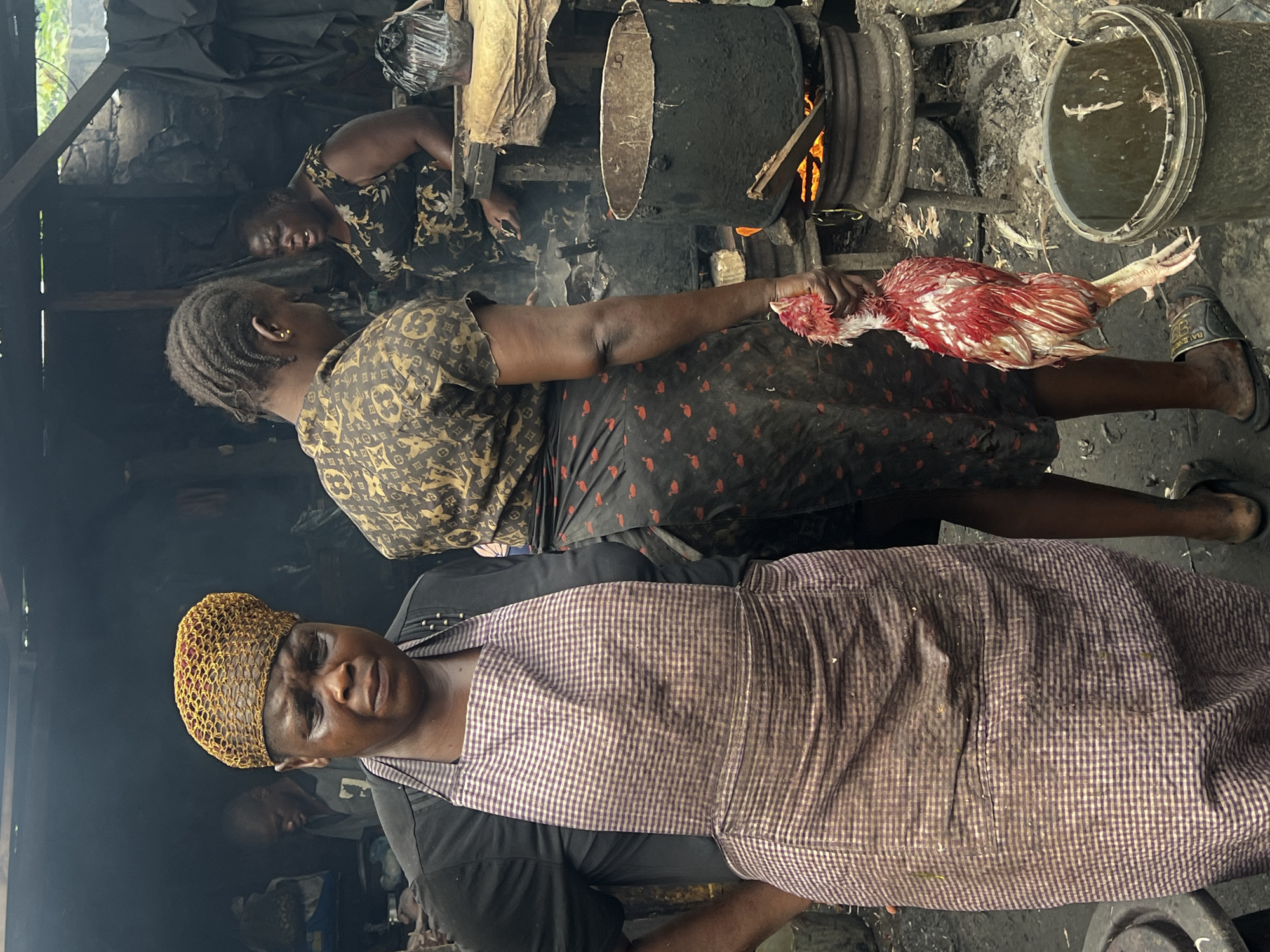 The image shows a busy outdoor market scene in Nkpor, Nigeria, likely a food stall or butchery. In the foreground, a woman wearing a black shirt, a checkered apron, and a yellow head wrap looks directly at the camera with a distressed expression. To her right, another woman in a patterned shirt and a skirt with a red flamingo print holds a raw, plucked chicken by its legs. Behind them, a large, dark pot sits over a fire, with steam or smoke rising. Another woman is visible in the background, tending to the cooking. The ground is dirt and appears wet and somewhat cluttered with debris. The environment is rustic and appears to be under a makeshift shelter.