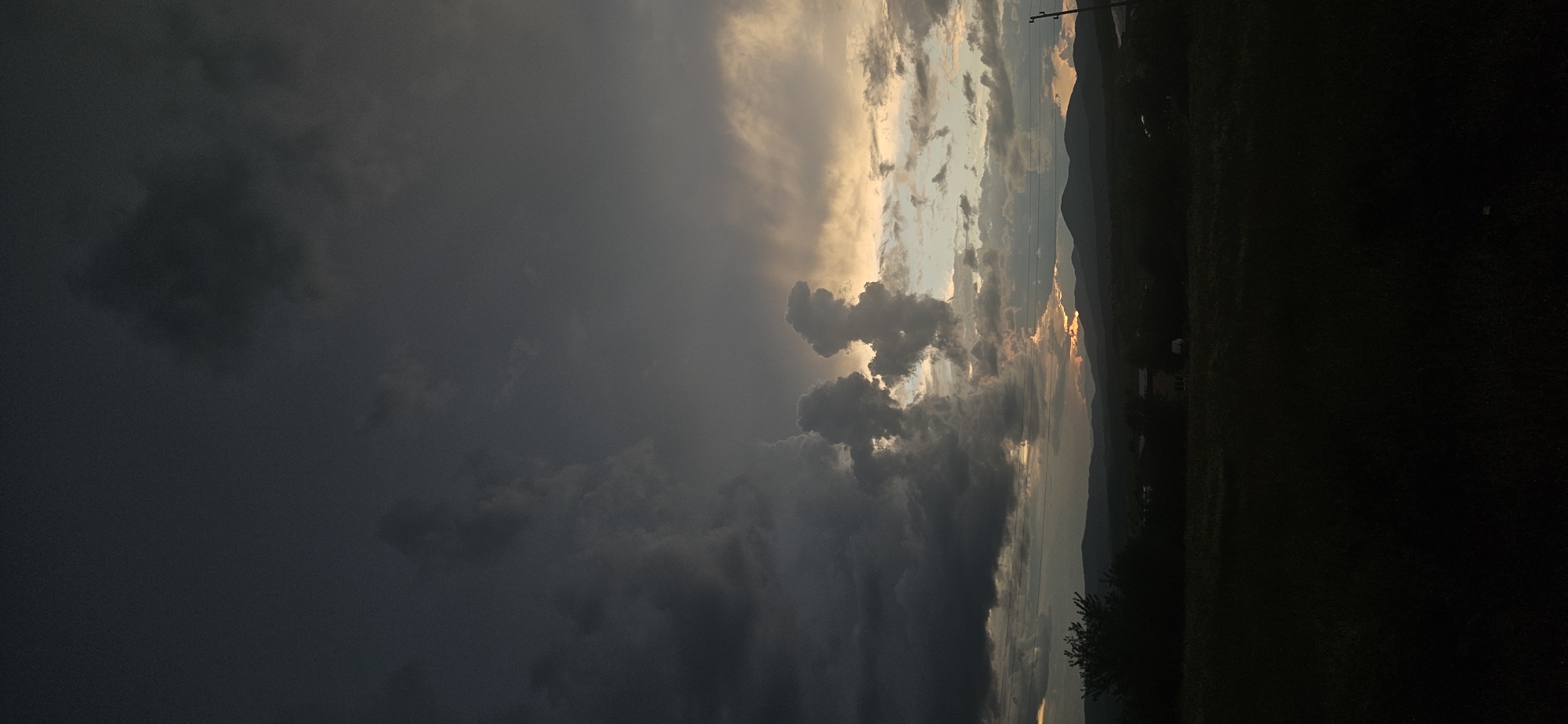 This image, captured in Momin Prohod, Bulgaria, presents a dramatic natural landscape during what appears to be either sunrise or sunset (golden hour). The scene is dominated by a dynamic sky. On the left side, dark, heavy storm clouds, deep gray to almost black, suggest dense atmospheric conditions or a recent downpour. Moving towards the center and right, the sky transitions to lighter hues, with vibrant golden, orange, and pale blue light breaking through the clouds. This light brilliantly illuminates the edges and undersides of sculpted cumulus-like clouds, creating striking high contrast and dramatic forms against the darker storm front.
In the background, a range of distant mountains is visible, largely silhouetted against the brighter portions of the sky, with warm colors hinting at the sun's low position on the horizon. A utility pole with power lines is present on the upper right, subtly indicating human presence within the natural vista. The foreground consists of dark, shadowy land, likely fields or forested areas, which is largely undifferentiated. No people or visible text are discernible in the image. The overall impression is one of powerful atmospheric change, showcasing the beauty and intensity of natural elements as a storm might be clearing or approaching.