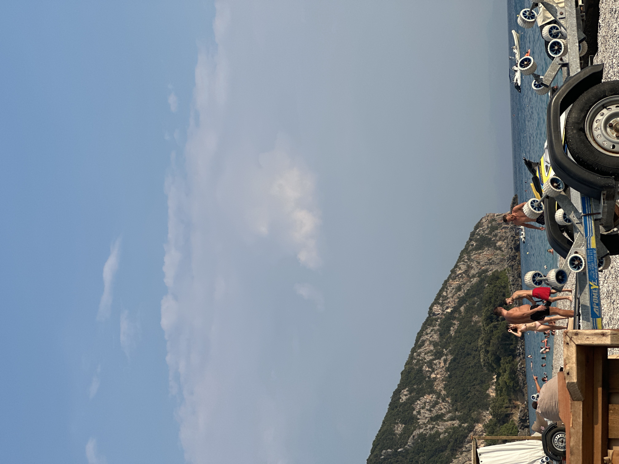 An eye-level, vertical outdoor shot captures a beach scene with a rocky, tree-covered mountainside in the background. The sky is a clear blue with scattered white clouds. In the foreground, a trailer with several wheels is parked on a pebble beach. A man in red shorts stands next to the trailer, talking to another man in black shorts. To the left of the trailer, a woman in a bikini stands with a man and another woman. In the water, several people are swimming and floating. A few boats are visible on the sea. The overall atmosphere suggests a bright, sunny day at a popular tourist destination.