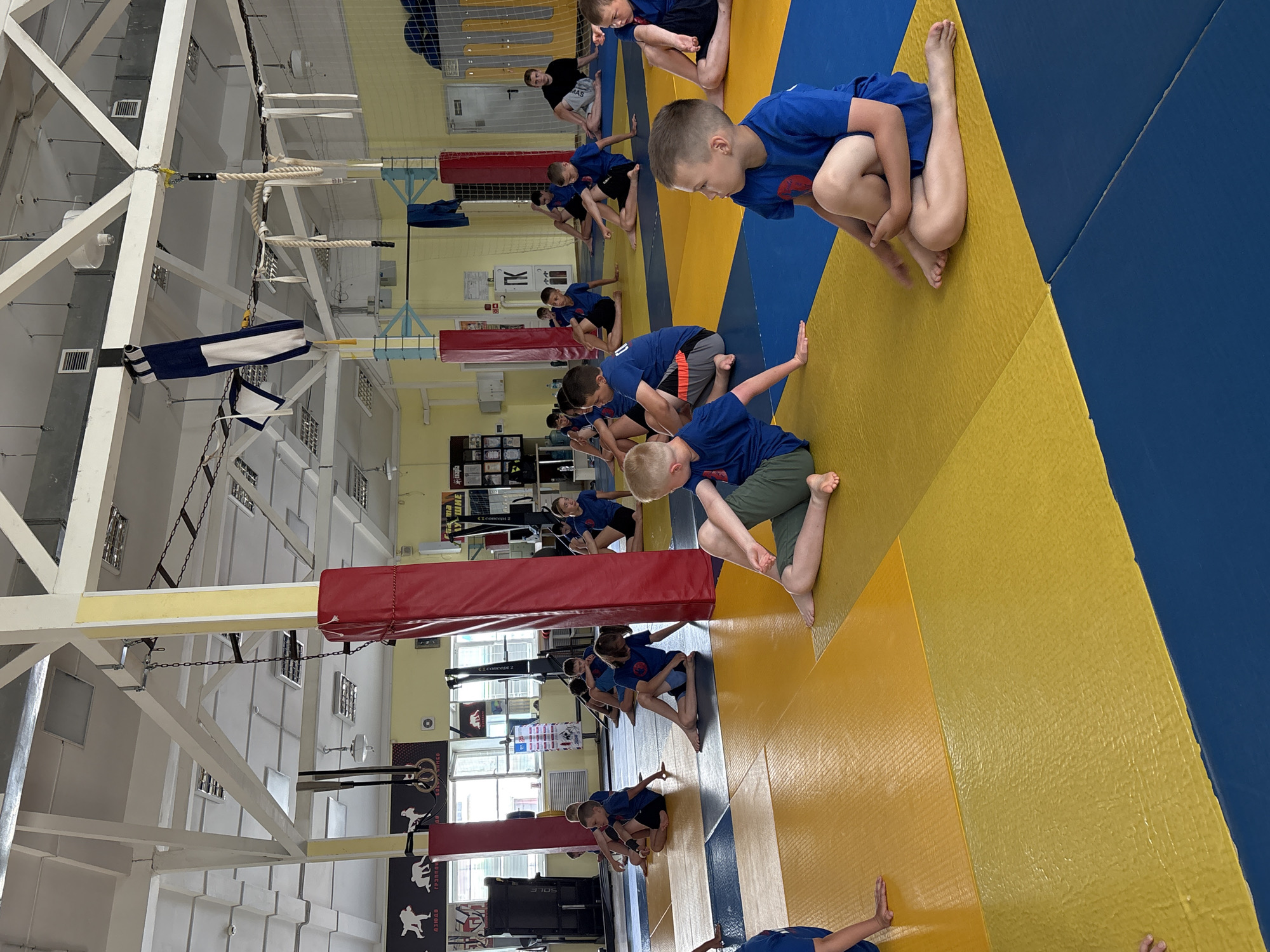 In a gymnasium in Chaikovsky, Russia, a group of young boys and girls, likely participants in a martial arts class, are seen performing stretching exercises on a blue and yellow matted floor. The room has a high, exposed ceiling with industrial lighting and exposed beams. Gymnastic equipment, including rings and ropes, are suspended from the ceiling. Posters and awards are displayed on the walls. The participants, dressed in blue t-shirts and shorts, are in various seated positions, demonstrating flexibility and focus. The scene suggests a training session in a sports facility.