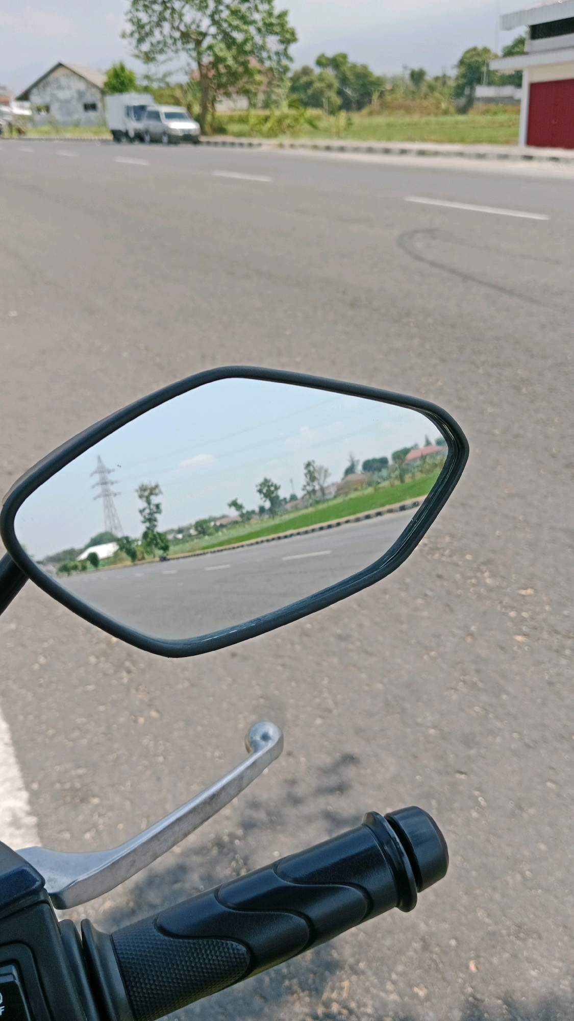 In Sukowinangun, Indonesia, the image captures a roadside scene from the perspective of a motorcycle. The foreground displays a motorcycle's black handlebar grip and a silver brake lever against an asphalt road surface. In the background, a reflective side mirror of the motorcycle shows a view of the road, a green field, houses, and trees. A prominent power pylon is visible in the distance on the left side of the mirror's reflection. The road appears to be a two-lane street with white lane markings. Along the right side of the road, a building with a red facade is partially visible. The environment suggests a sunny day with a clear sky.