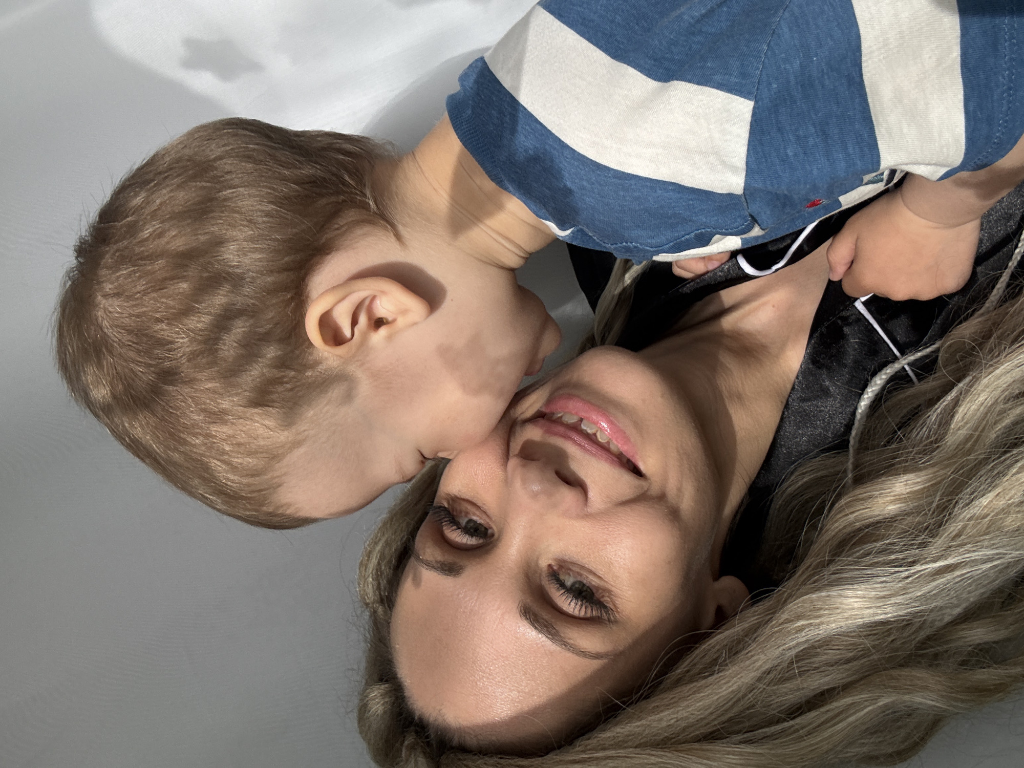 A close-up shot shows a woman and a toddler in an intimate moment. The woman, who has fair skin, long blonde hair, and prominent eyelashes, is seen smiling slightly. A toddler, who is wearing a blue and white striped shirt, appears to be kissing her cheek. The background is a plain, light-colored surface, possibly a wall or floor. The location and city are Unknown, Unknown.