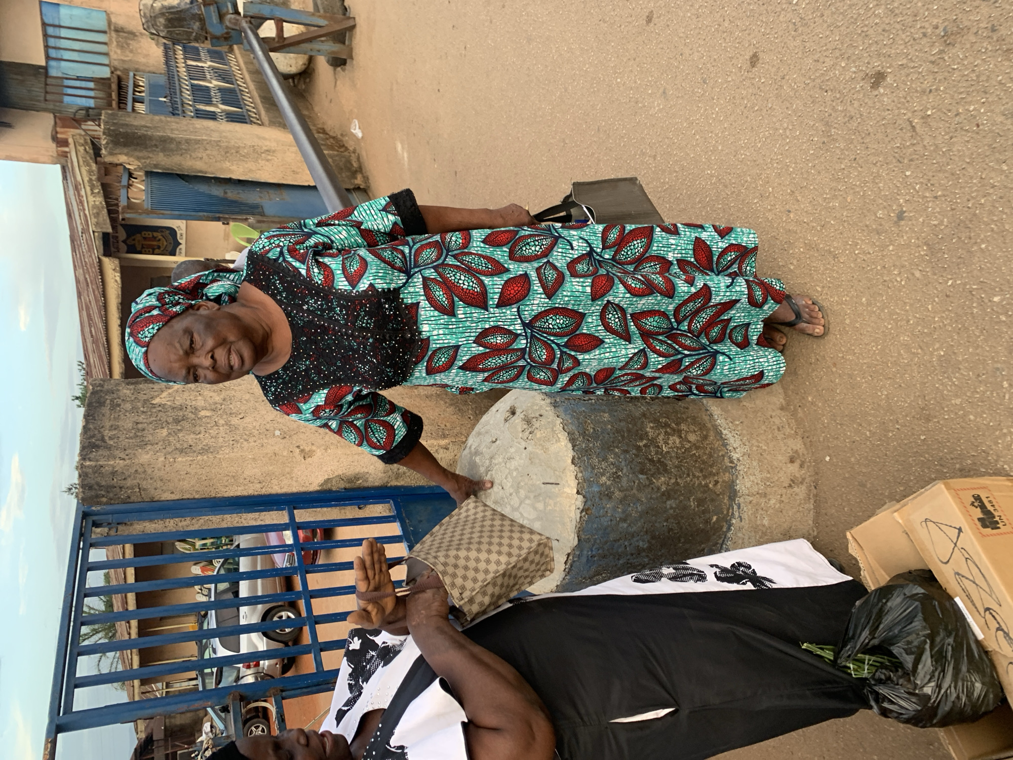 This outdoor image from Jos, Nigeria, features two women on what appears to be a street or pathway, with buildings and a blue metal gate in the background.
The primary subject is an older woman standing in the foreground, looking directly at the camera with a gentle smile. She is dressed in a vibrant, full-length garment and a matching headwrap, both made of a patterned fabric featuring green, red, and white leaf-like designs on a black background, typical of African print (Ankara). She wears simple dark flip-flops and holds a small dark bag.
Beside her, a second woman is partially visible, appearing to be sitting or leaning. She wears a black and white patterned top and a black skirt or wrapper. Her right hand is visible, open. Between the two women, a light-colored, checkered handbag rests on a large, cylindrical concrete block or pedestal.
The ground is a light brown, rough-textured surface. In the background, a blue metal gate provides a glimpse into a property where several cars are parked. Beyond the gate, concrete buildings with blue window shutters are visible, and a long metal pole extends upwards from one structure. Near the seated woman, cardboard boxes and a black plastic bag containing green leafy items are on the ground. Text "UN 3481" and "DANGEROUS GOODS" is partially visible on one of the boxes.
The scene is lit by soft daylight, suggesting late afternoon. The overall setting is a casual, everyday street environment.