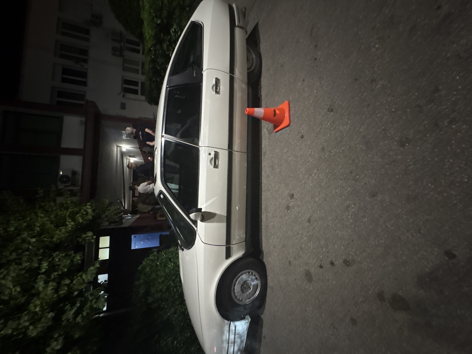 The image shows a white car parked on a street in Podgorica, Montenegro, based on the building style. An orange traffic cone is positioned next to the car. In the background, people are visible near a building. Foliage flanks the left side of the frame, and the street appears to be asphalt. The lighting suggests it is evening or night.