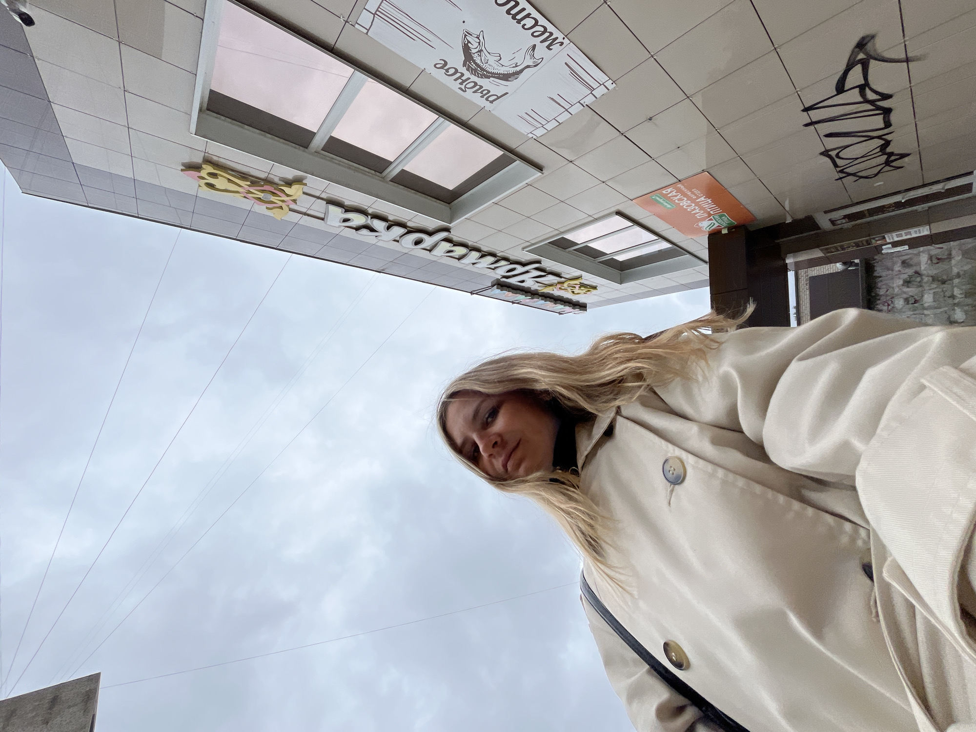 The image shows a woman from a low-angle perspective, looking up at the camera. She has blonde hair and is wearing a light-colored trench coat. Behind her, a building with beige tiled walls is visible. A sign on the building reads "Зомарка" (Zomarka). Another sign on the building displays a drawing of a fish and the words "рыбное место" (fish place). To the right of these signs, there is graffiti on the lower part of the building wall. The sky above is overcast. The building appears to be a commercial establishment. The signage suggests the location is in Chaikovsky, Russia, as "Зомарка" is a known supermarket chain in the region and "Глазовская птица" (Glazovskaya ptitsa), also visible on another orange sign, is a poultry producer often found in such locations.