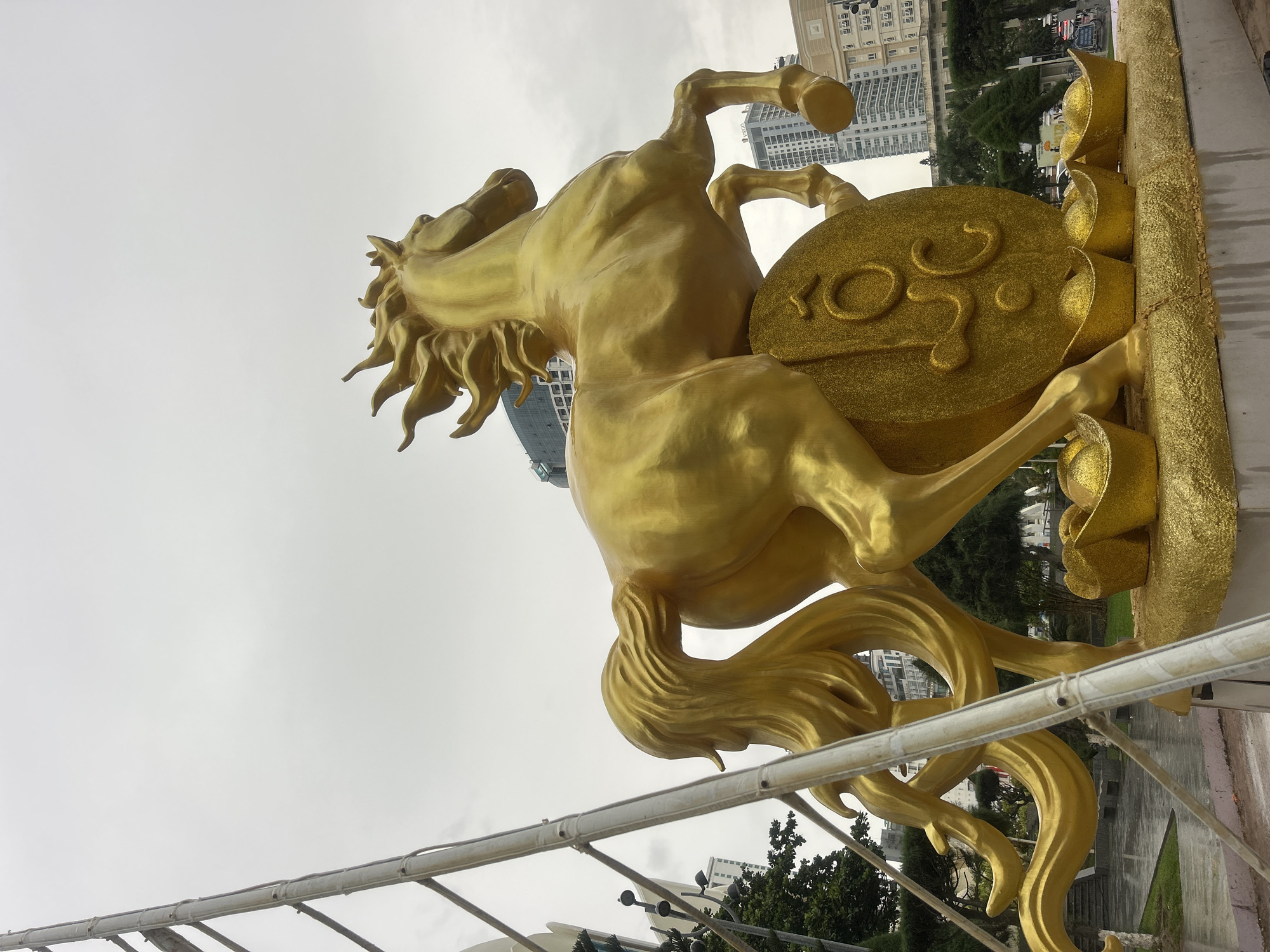 A low-angle shot depicts a golden horse statue adorned with Chinese gold ingots, set against an overcast sky. The statue, the centerpiece of the scene, is located in Nha Trang Ward, Vietnam, as evidenced by the architecture and vegetation in the background. The horse is positioned as if rearing, and it is decorated with a large golden disc featuring embossed Chinese symbols. The base is ornamented with additional gold ingots, a typical symbol of prosperity and good fortune.
The buildings in the background feature modern architecture, suggesting an urban setting. The landscaping includes trees and greenery typical of Southeast Asia. The presence of a metal structure indicates that the statue may be part of an outdoor public display.