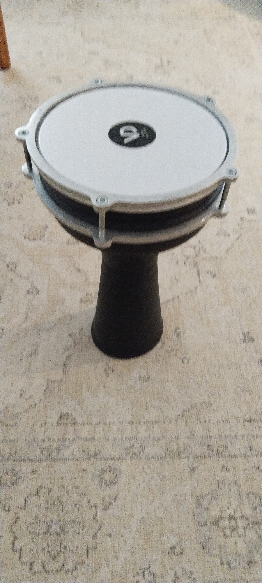 The image shows a darbukka drum, a Middle Eastern percussion instrument, placed on a patterned rug. The drum is black with a white drumhead, featuring a dark oval logo with the letters "VD" in white. The rug has a light-colored base with a faded, intricate pattern in darker shades. The scene appears to be indoors, possibly in a home, as suggested by the domestic setting of the rug and the lighting. No specific location in Buca, Turkey, can be identified from the image alone.