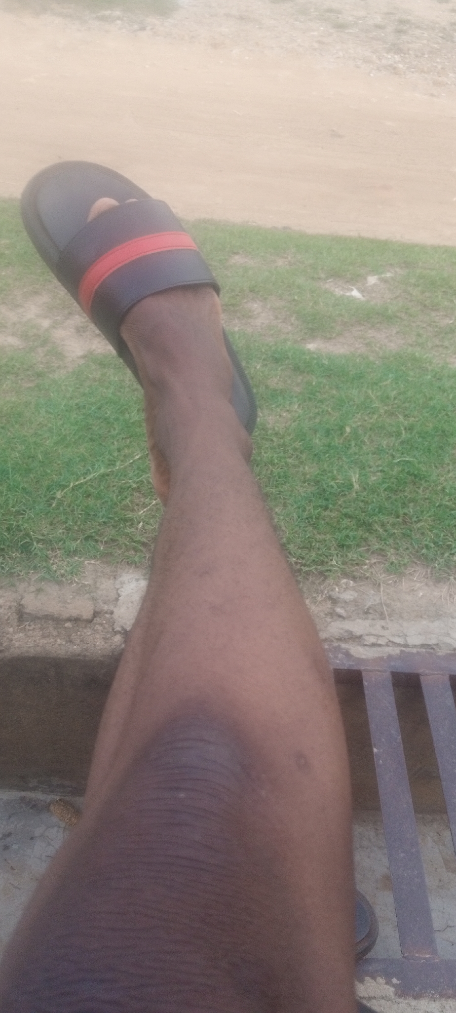 A close-up, eye-level shot shows a dark-skinned leg extended forward, wearing a black sandal with a red stripe. The leg rests on a metal grate, with a concrete edge visible behind it. In the background, there is grass and what appears to be bare ground. Based on the location of Narayi, Nigeria, there is no evidence to show if the location is in Narayi Nigeria.