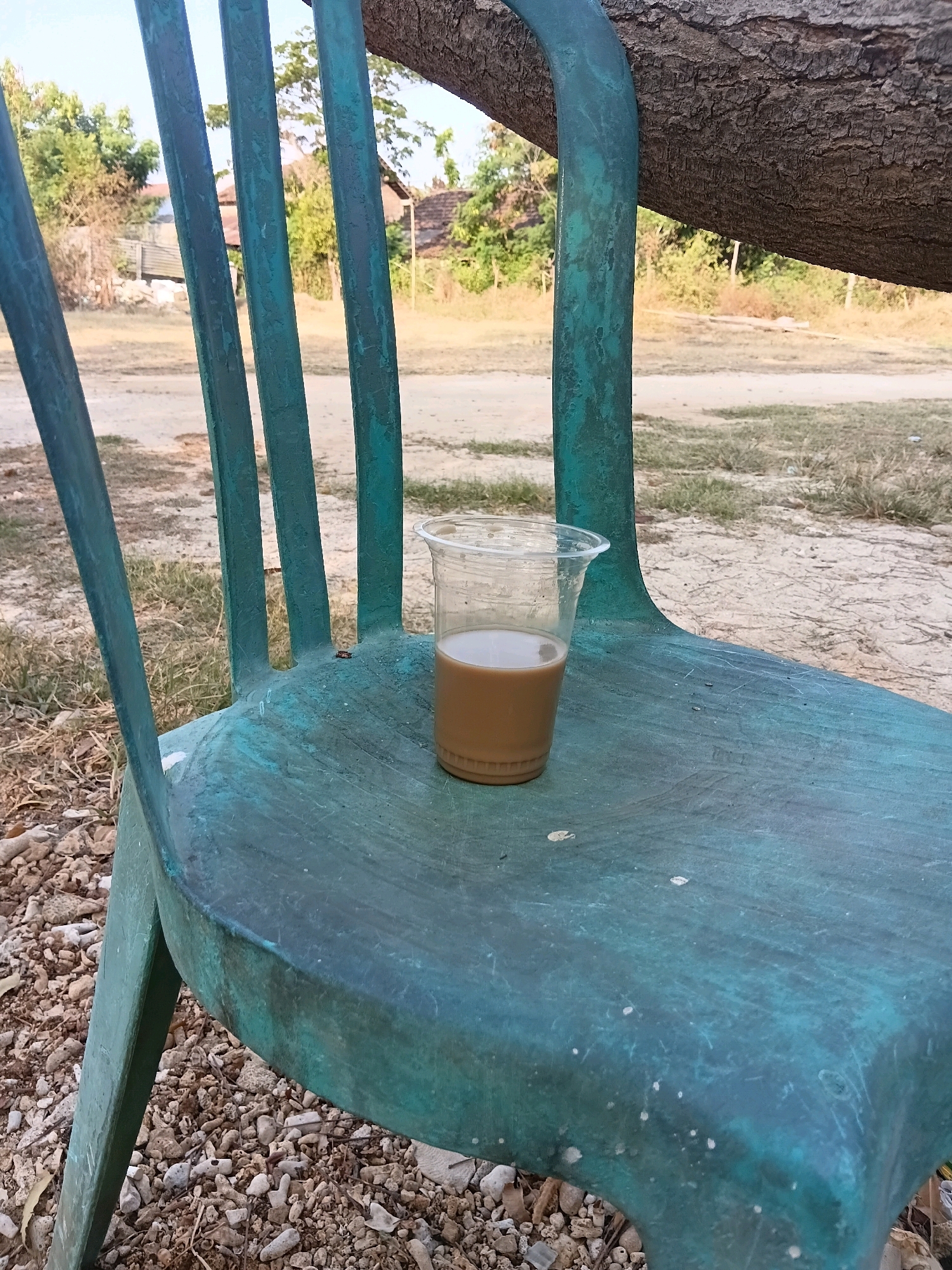 A plastic cup filled with a light brown liquid sits on the seat of a teal-colored metal chair. The chair is positioned outdoors on a ground covered with small stones and dried leaves. In the background, a tree trunk arches over the scene, and beyond that, there is a dirt road, sparse vegetation, and a few buildings, suggesting a rural or semi-rural setting. The sky is clear and bright. The overall impression is one of a still life, with the focus on the cup and chair in a natural environment. The image appears to have been taken in Cikal, Indonesia.