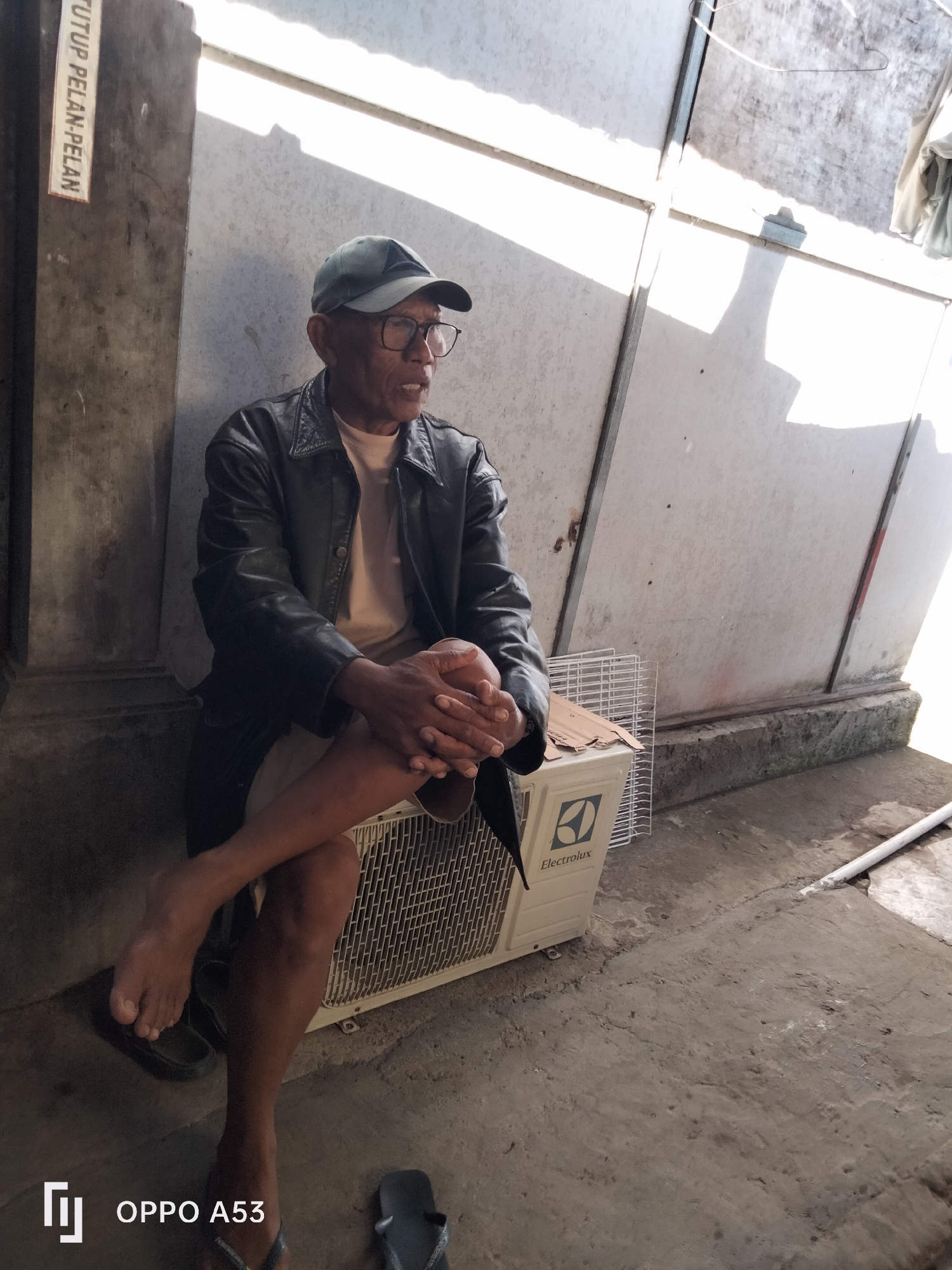 An elderly Asian man, wearing a dark leather jacket, a light-colored t-shirt, and a baseball cap, sits with his legs crossed on an air conditioning unit outside. He is wearing glasses and his hands are clasped. The setting appears to be an outdoor area with a concrete floor and a weathered wall behind him, with bright sunlight casting shadows. A sign with Indonesian text, "TUTUP PELAN-PELAN" (close slowly), is visible on the wall to the left. An Electrolux air conditioning unit is beneath him, with a white wire rack on top. Flip-flops are visible on the floor near his feet. The overall scene is one of a person resting in a somewhat utilitarian outdoor space.