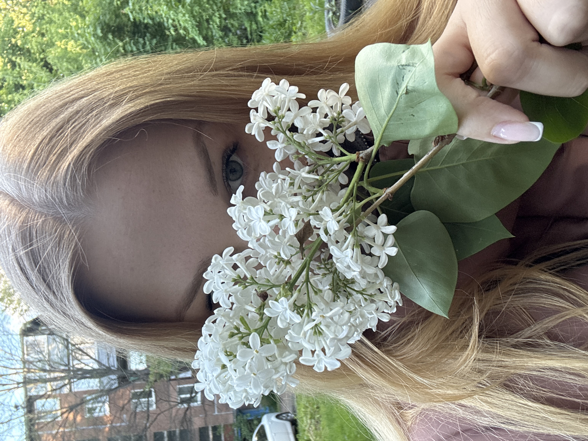 A fair-skinned woman with blond hair is holding a branch of white lilacs in front of her face. One of her eyes is visible behind the flowers. She has manicured nails with white tips. The background is blurry but appears to show trees, a building and a white car. I am unable to verify that this image was taken in Rusanovo, Russia, as I am not able to geolocate the image.