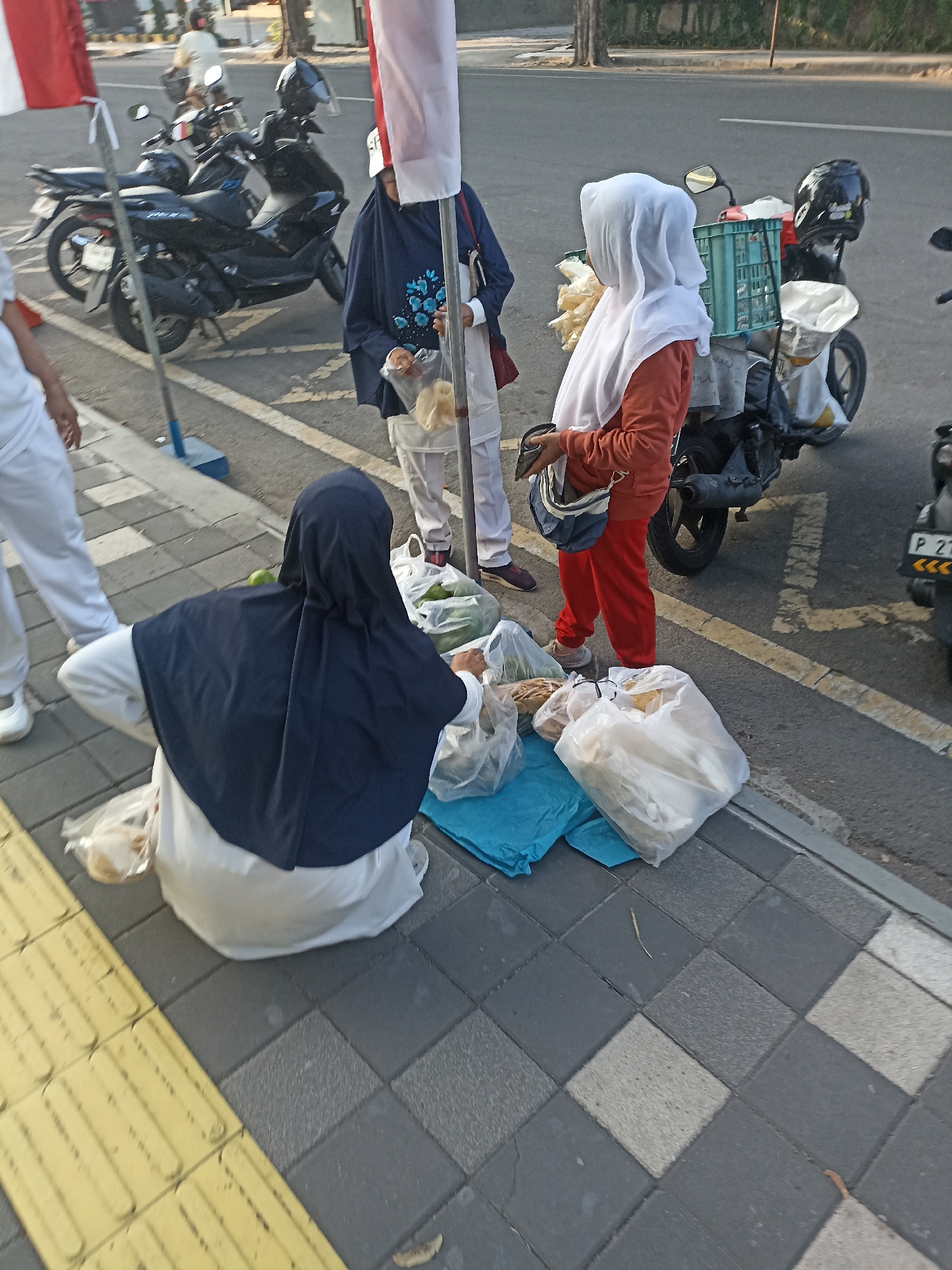 In Kelurahan Kepatihan, Indonesia, a sidewalk scene unfolds with three individuals and several motorcycles. One person, facing away from the camera and wearing a dark blue hijab and white top, is crouched. To their right, a person in a white hijab and orange top is standing. Another individual, mostly out of frame on the left, is wearing white pants. Two motorcycles are parked behind the standing individuals, one with a blue crate on the back. Various plastic bags containing what appear to be food items are scattered on the sidewalk and on the ground near the crouched person. The environment is a paved sidewalk adjacent to a road with yellow and white markings, suggesting a public street.
