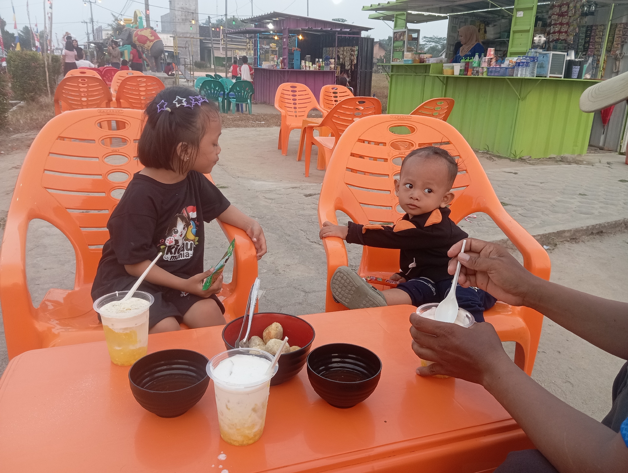 In Way Dente, Indonesia, two young children, a girl and a boy, sit at an orange plastic table outside a food stall. The girl, wearing a black t-shirt with a cartoon print and a purple star-shaped hair accessory, looks towards the boy. The boy, in a black shirt with orange accents, gazes to his right. On the table, there are several plastic cups containing drinks, one with a spoon, and two black bowls, one holding what appears to be fried snacks. An adult's hands are visible, holding a spoon in a plastic cup of drink. In the background, other orange and green plastic chairs are arranged, and various stalls and structures are visible, suggesting a market or outdoor dining area. The ground is covered in gravel.