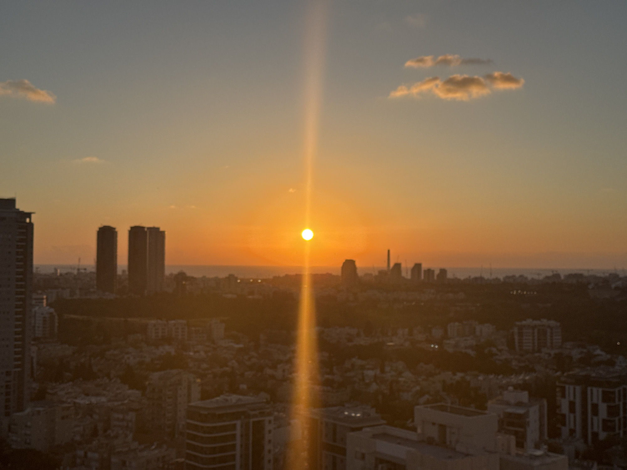 The image shows a cityscape at sunset in Ramat Gan, Israel. The sun is setting on the horizon, casting a strong vertical glare through the center of the frame. The buildings in the foreground are densely packed, with a mix of high-rise and low-rise structures. The skyline is punctuated by taller buildings, including three prominent skyscrapers. The color palette is dominated by warm tones of orange, yellow, and tan. The sea can be seen on the horizon. There are a few clouds in the sky.
