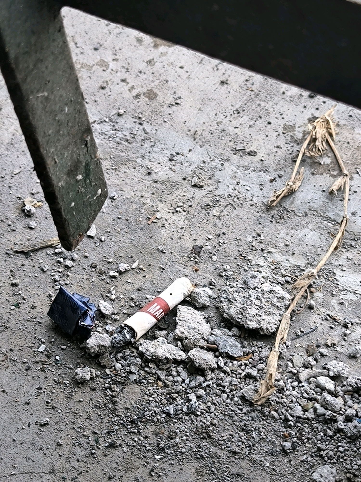 A close-up, high-angle shot shows a cigarette butt and a small dark object on a rough, grey, concrete surface. The cigarette butt is white with a red band that has the letters "MAR" printed on it, and it appears to have been smoked. To the left of the cigarette butt is a small, dark, rectangular object wrapped in shiny, crinkled material, possibly tape. A thin, dried plant stalk lies in the upper right portion of the image, extending from the top edge downwards, with its delicate, skeletal form contrasting with the rough concrete. A dark, angled metal object is visible in the upper left corner, casting a shadow. The concrete surface is littered with small pebbles and debris.