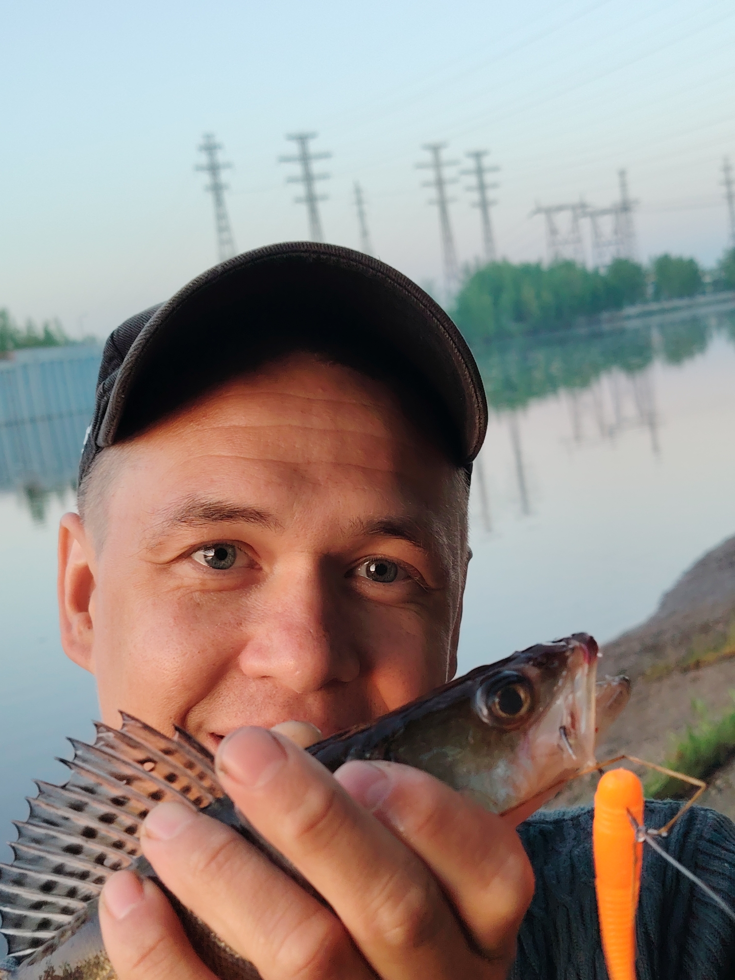 A close-up shot shows a man holding a fish, obscuring part of his face with it. The man has short hair and blue eyes, and wears a dark cap. The fish is light-colored with distinct dark spots on its dorsal fin, and an orange lure attached to its mouth. In the background, there is a body of water reflecting the sky, and structures that resemble power lines are visible. The location is identified as the city of Chaikovsky, Russia.