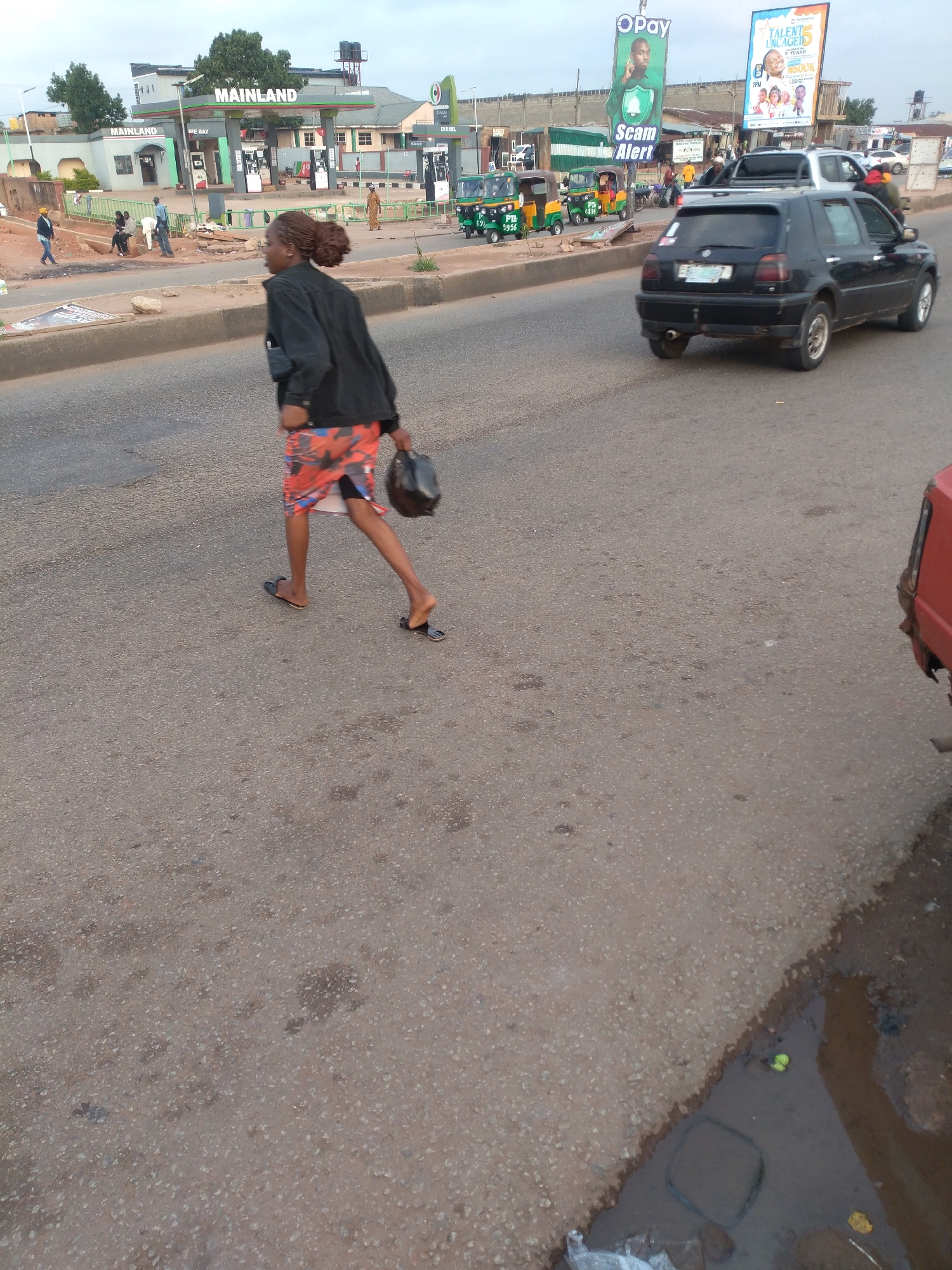 The image captures a street scene in Gura Top, Nigeria, focusing on a woman crossing the road. She is wearing a black jacket and a colorful patterned skirt, with black sandals on her feet. In her right hand, she carries a black bag. She is walking from left to right across the asphalt road, which shows signs of wear and tear.
In the background, a "MAINLAND" gas station is visible with several fuel pumps. Other vehicles, including a black Volkswagen Golf, a white pickup truck, and several brightly colored auto-rickshaws (Keke Napeps), are present on the road and sidewalk. A red vehicle is partially visible in the lower right corner of the frame. Advertisements, including one for "Scam Alert," are displayed on billboards. Several other individuals are seen in the background near the gas station and on the roadside, some appearing to be working or interacting. The environment suggests a typical commercial and transportation hub within the city.