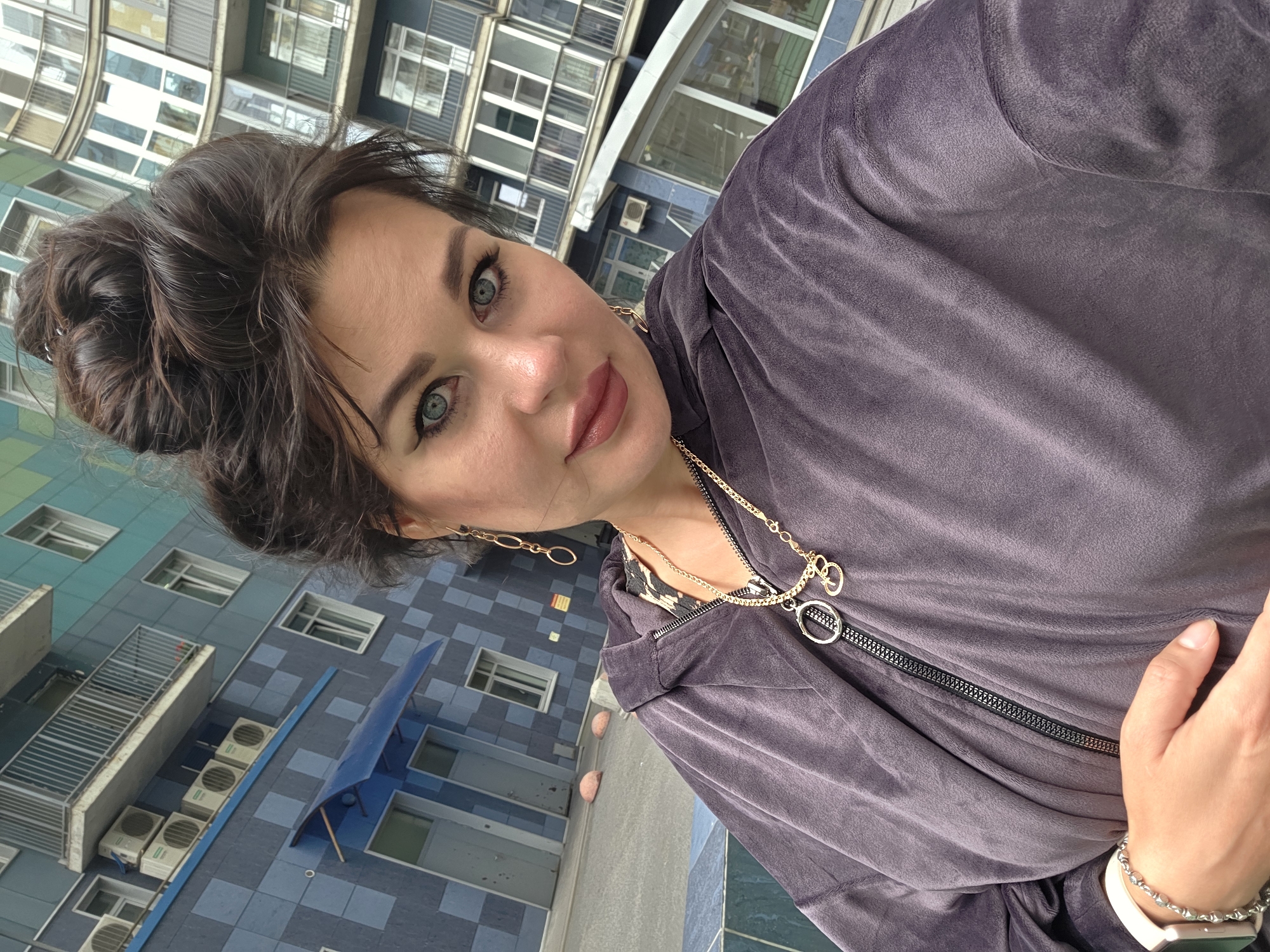 A woman with dark hair styled in a bun takes a selfie outdoors in front of a modern apartment building with a blue and teal facade. She is wearing a dark purple velour hooded jacket, a gold necklace with circular and numerical charms, and gold dangling earrings. Her makeup includes winged eyeliner and lip gloss. Her left hand rests on her chest, and she is wearing a white smartwatch with a silver band on her left wrist. The background shows several balconies and air conditioning units attached to the building. The ground is paved with asphalt and there are two pink bollards visible. The building's design suggests it is located in a contemporary urban residential area.