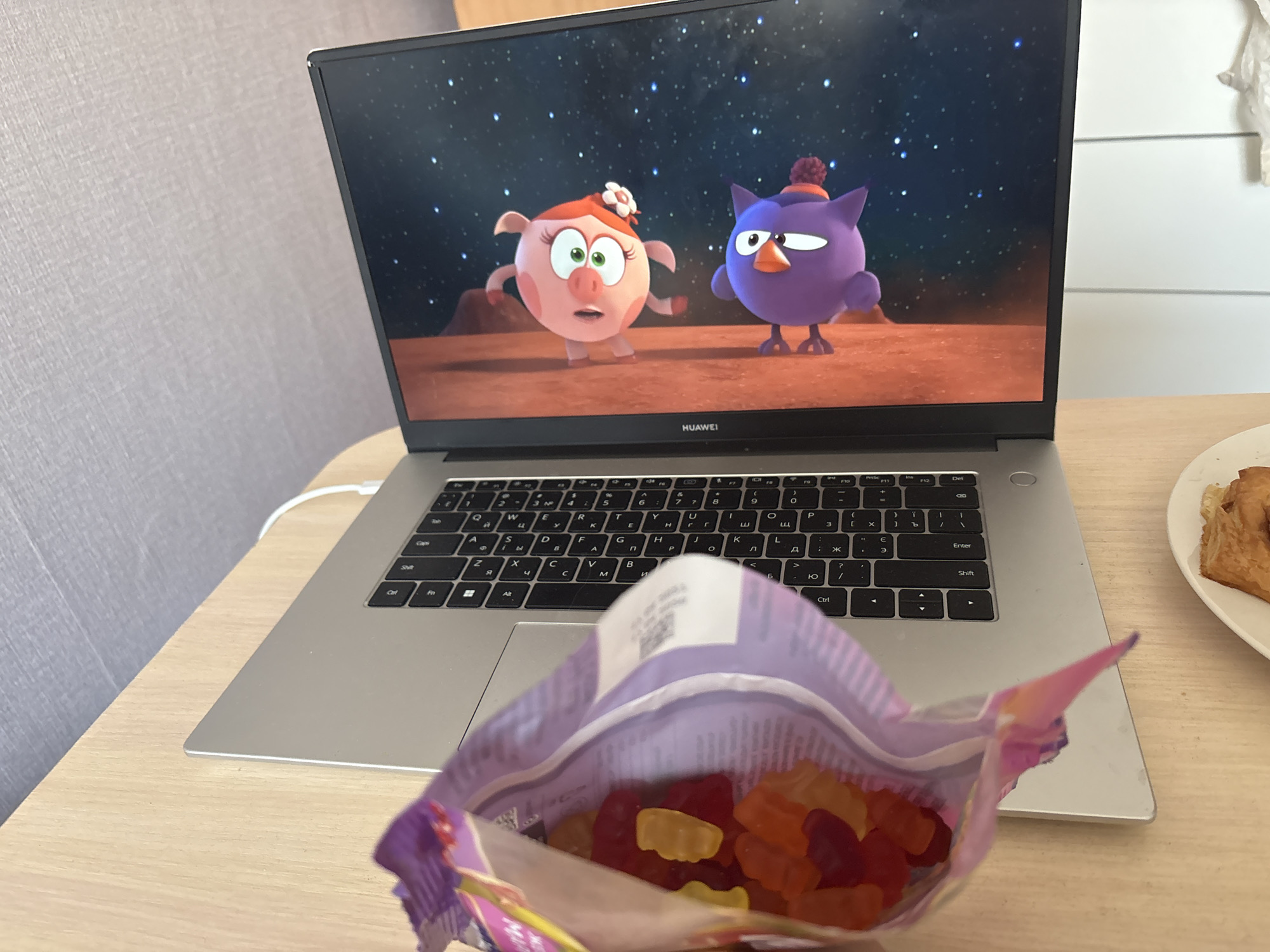 The image depicts a laptop on a light-colored surface, likely a table. On the screen, an animated show is playing with two cartoon characters: a pink pig with a white flower on its head and a purple bird wearing a matching purple hat. In front of the laptop, there's an open bag of gummy bears. To the right of the laptop is a white plate with pastries. The laptop itself appears to be a Huawei model. The background is a plain wall.