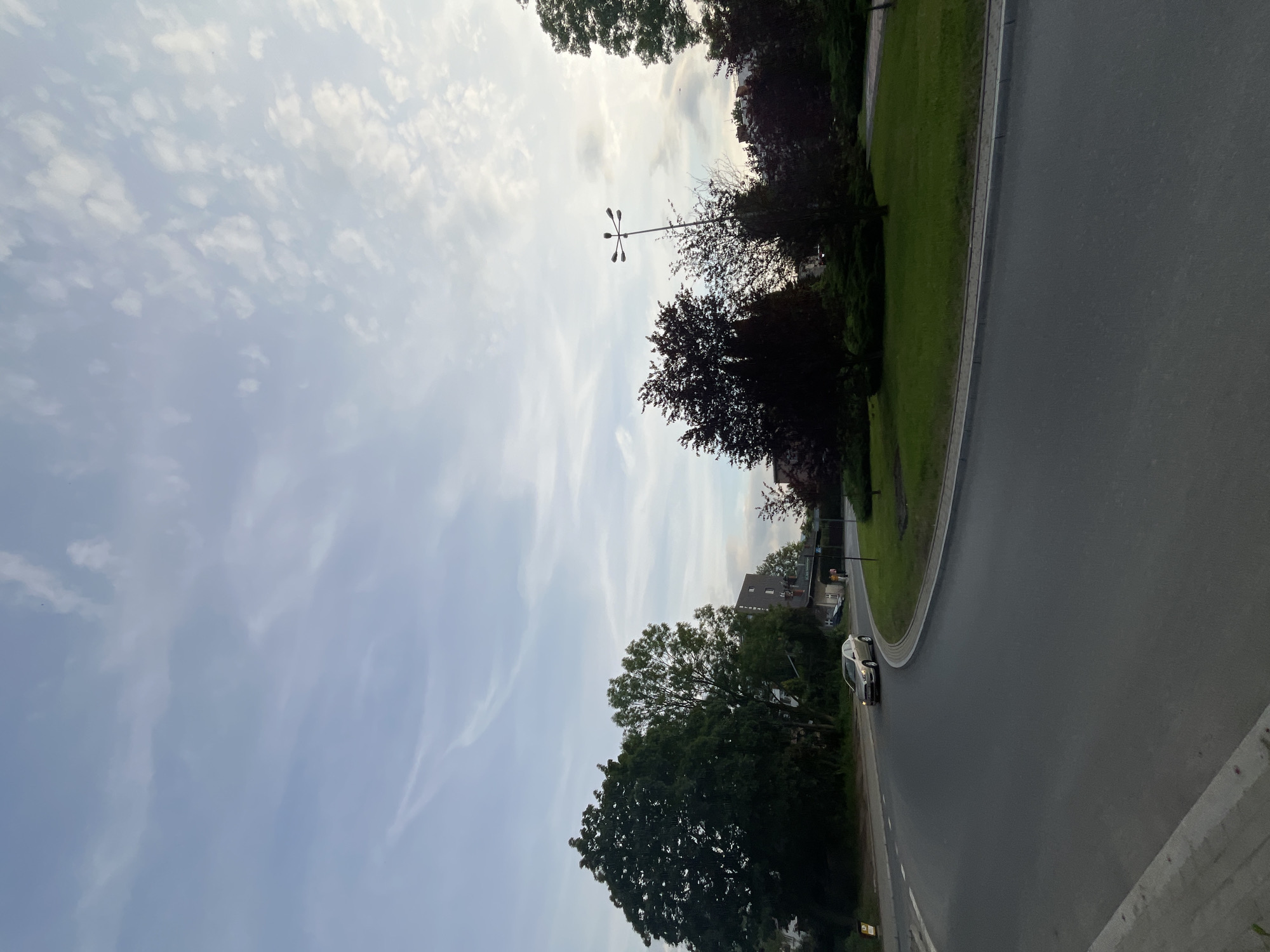 The image captures an outdoor scene dominated by a vast sky, a curving road, and various trees. The perspective is slightly elevated, looking over a road and a green area.
**Subjects:**
The upper and left portions of the image are filled with a light blue sky, adorned with scattered, wispy white clouds that stretch across the frame, indicating cirrus cloud formations. Below the sky, a varied line of trees forms a significant visual element. Some trees display vibrant green leaves, while others exhibit darker, possibly reddish-brown or deep green foliage. Centered against the sky and trees, a modern street light with multiple (specifically, five) lamp heads extends upwards.
To the right and bottom of the image, a dark asphalt road curves around a grassy median. A thin white line marks the edge of the road. A light-colored sedan, appearing silver or champagne, is visible on the road, driving away from the viewer along the curve. In the background, partially obscured by trees, a building with a dark roof and light-colored walls can be seen. In the immediate bottom-right foreground, a segment of a concrete barrier or wall is visible.
**Activity or event taking place:**
A car is traveling along the curving road. No other significant activity is depicted.
**Notable details:**
The time of day appears to be late afternoon or early evening, evidenced by the soft, diffused lighting and the absence of harsh shadows, suggesting the sun is low or behind clouds. The weather is partly cloudy and bright. The arrangement of the curving road, green spaces, trees, and a street light suggests a well-maintained public area, possibly a boulevard, park-like street, or a road leading to a roundabout within a suburban or urban environment. There is no visible text in the image.