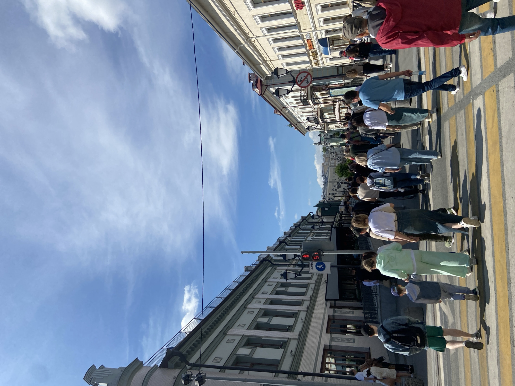 The image depicts a bustling pedestrian street in Kazan, Russia, under a clear blue sky with scattered white clouds. Approximately 30-40 individuals, predominantly adults and some children, are seen walking away from the camera, primarily across a prominent zebra crossing.
On the left, a multi-story building features a light-colored ground floor with large windows and darker upper floors with regularly spaced rectangular windows. A distinctive, ornate, turret-like structure is visible at the building's corner in the upper-left, and a dark railing runs along its rooftop. On the right, similar multi-story buildings, primarily cream-colored, display classical architectural elements such as framed windows, cornices, and some balconies. Storefronts are visible at street level on both sides of the street.
Street infrastructure includes a red pedestrian traffic light displaying "5" seconds remaining, positioned near a sign reading "ЗОНА" (ZONE) with a pedestrian symbol, confirming a pedestrian area. Several ornate black street lamps line both sides of the street, extending into the distance. A circular sign with a red diagonal line through a scooter icon indicates a prohibition on scooters. A single dark cable horizontally traverses the upper-middle section of the frame.
Pedestrians are dressed in casual attire, consistent with warm weather. Notable individuals in the foreground include a young man with a backpack and green shorts, a child in a grey tracksuit holding the hand of a woman in a mint green tracksuit, a woman in a long denim skirt, and a man in a blue t-shirt carrying a plastic water bottle. Shadows cast by the people and buildings indicate strong sunlight.