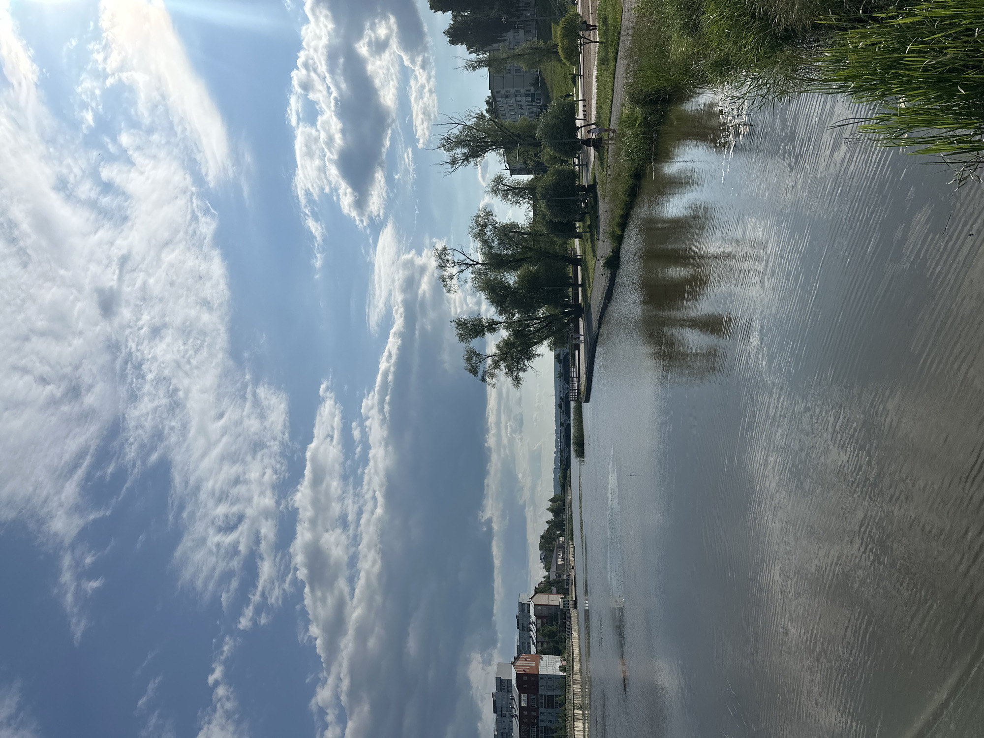 This is a vertical, full-shot photograph taken on a sunny day in Votkinsk, Russia. The image depicts a canal or river with a walkway running alongside it. The sky is a bright blue, filled with scattered white and grey clouds, with the sun shining through from the top left, creating a lens flare effect. The water reflects the sky and the surrounding greenery and buildings.
On the walkway, several people are visible. Some appear to be walking or strolling, while others are engaged in conversations or standing. There are trees lining the walkway, providing shade and enhancing the natural beauty of the scene. Behind the trees, residential buildings and some taller structures can be seen, indicating an urban or semi-urban setting. The overall atmosphere seems peaceful and relaxed, suggesting a leisurely afternoon.
The time of day is likely midday or early afternoon due to the bright sunlight and the shadows cast. The weather is pleasant, with clear skies and clouds. The location cues, such as the architecture of the buildings and the style of the walkway, are consistent with a Russian setting. There is no visible text in the image. The image captures a serene moment in a scenic waterfront area, popular for recreation and relaxation.
