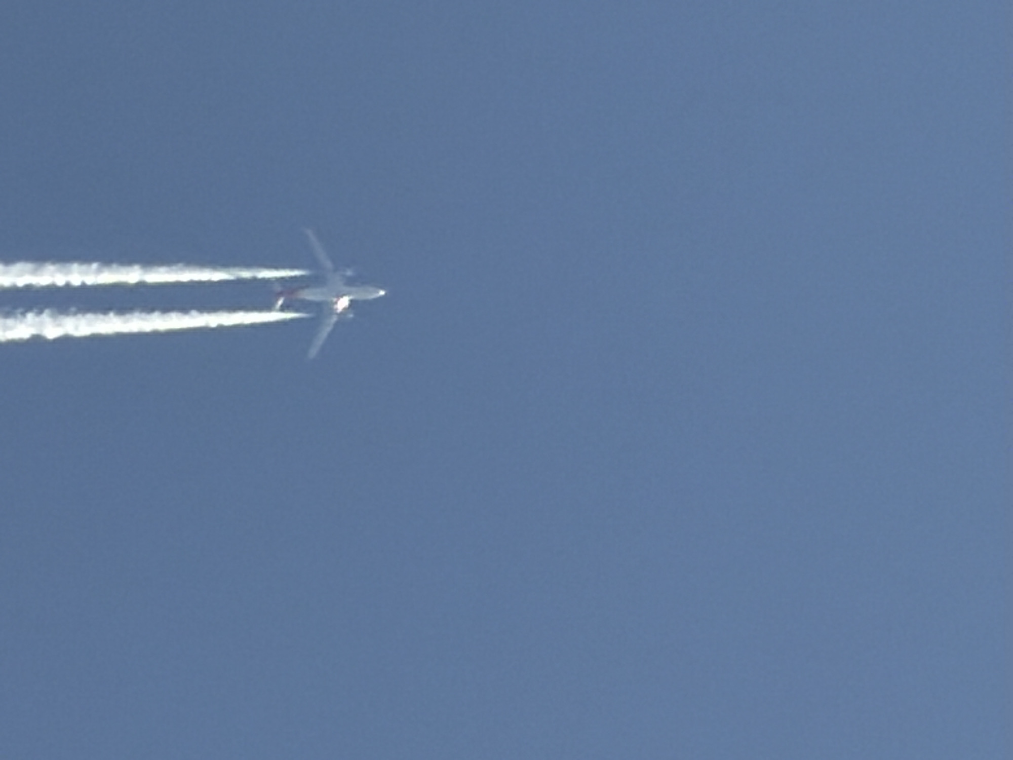 The image shows an airplane flying high in the sky. The plane is leaving a white contrail behind it. The sky is clear and blue. The location is not identified.