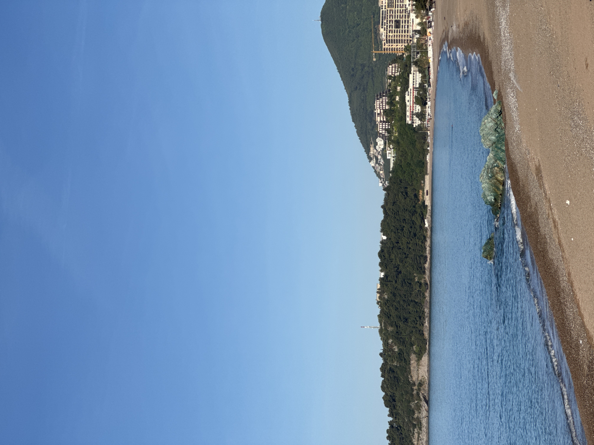 The image shows a coastal scene in Rafailovići, Montenegro. The right side of the image depicts a sandy beach meeting the blue water. The coastline is lined with a densely vegetated area transitioning into urban development with residential buildings and a construction site with a crane visible. A mountain backdrop further defines the scene. The left side of the image is dominated by a clear blue sky.