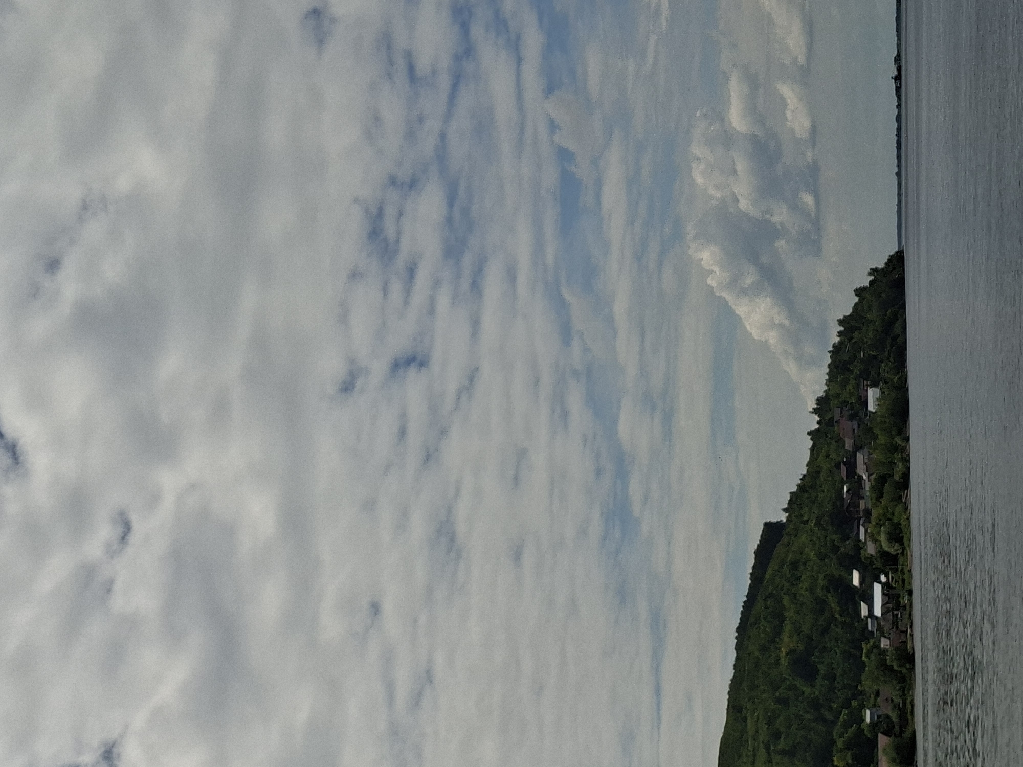 The image depicts a wide body of water in the foreground, likely a river, with gentle ripples on its surface. In the mid-ground, a densely forested hillside slopes down to the water's edge. A small village or settlement is nestled at the base of the hill, characterized by numerous houses with visible roofs. The vegetation is lush and green, covering the entire hillside. The sky above is filled with a mix of clouds; some are thin and wispy, creating a dappled effect, while others are larger and more voluminous, appearing as puffy white formations. The overall scene suggests a tranquil, rural landscape. This scene is consistent with the appearance of the Volga River near Verkhny Uslon, Russia, a region known for its scenic riverbanks and forested hills.