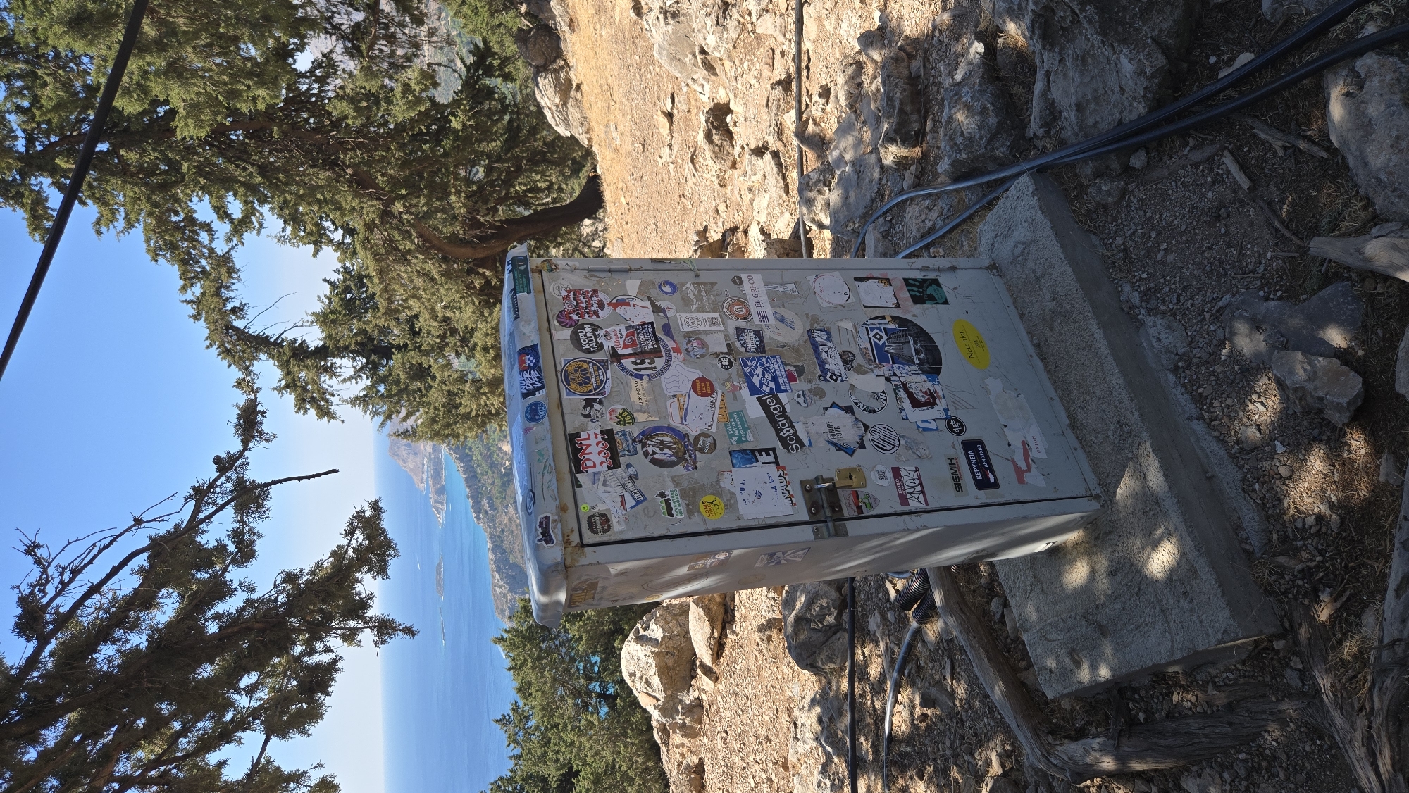 A medium shot captures a metal utility box covered in various stickers, placed on a concrete base in a rocky, outdoor environment. The box is gray and shows signs of wear. It is situated on a hillside with sparse vegetation and rocks. In the background, a body of water, possibly the Mediterranean Sea, is visible, with a coastline featuring hills and trees. The sky is clear and blue. The scene is illuminated by sunlight, casting shadows. The location is identified as Kolymbia, Greece.