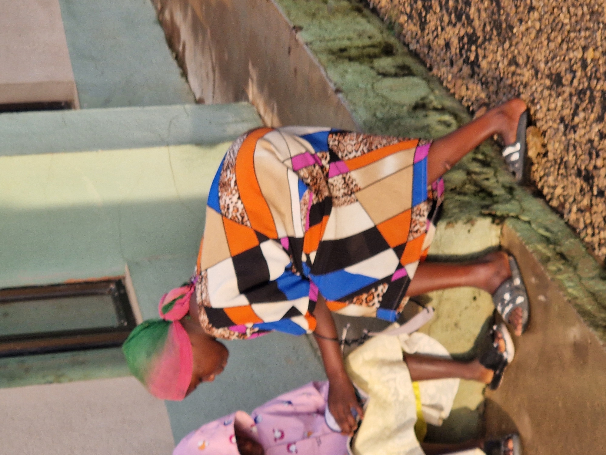 In an outdoor setting, two young girls are interacting with each other, one on the left wearing a pink hooded jacket with rainbow motifs, and the other on the right wearing a brightly colored, patterned dress. The girl on the left is seated and appears to be holding a book or notebook. The girl on the right is kneeling beside her and looking down. They are both wearing sandals. The environment appears to be a concrete walkway with a rough, textured curb or ledge running along the side, covered in small stones. Behind them, a light green, textured wall is visible with a dark-framed window. The overall lighting suggests it is daytime, with some shadows cast on the ground.