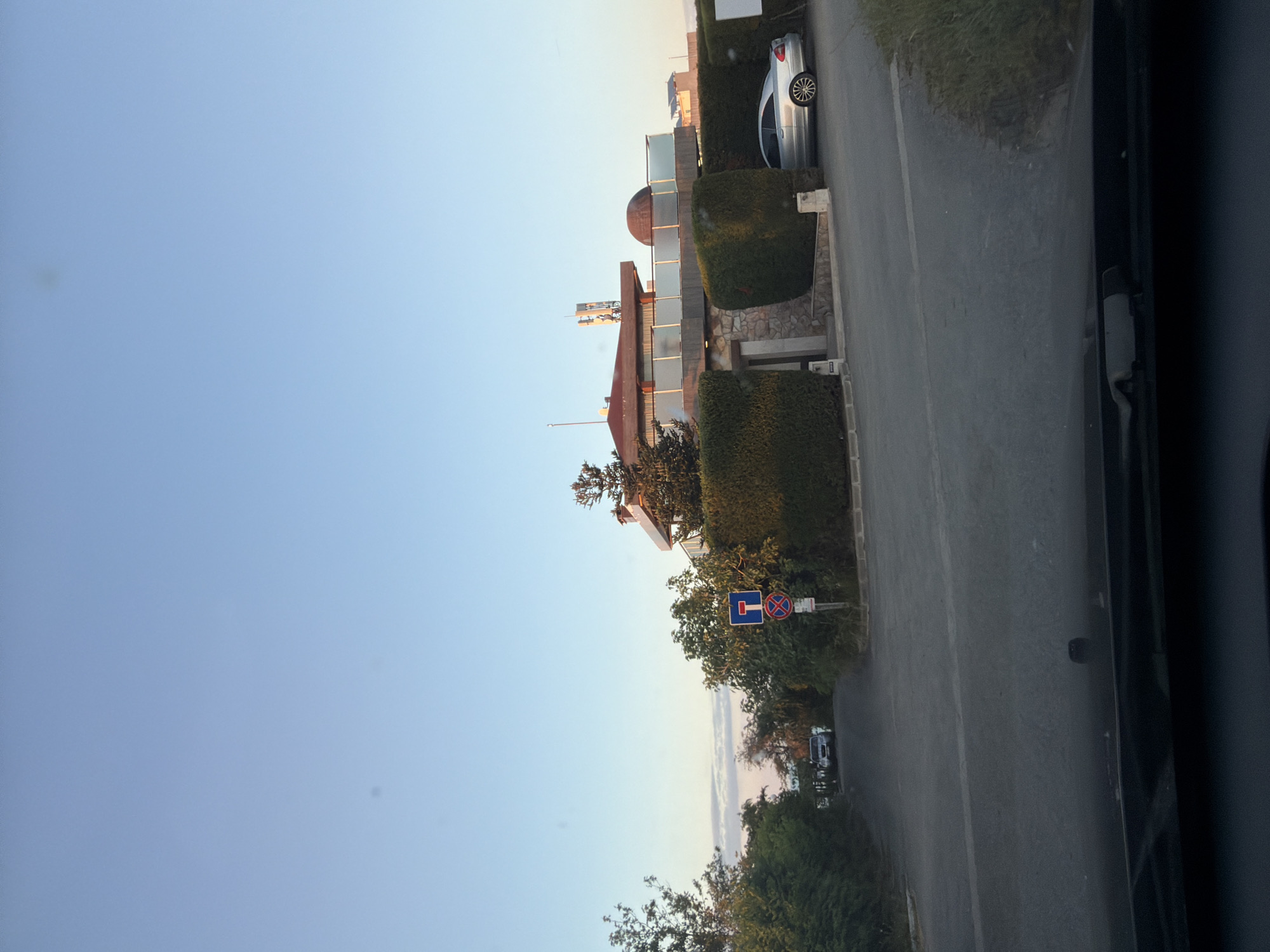 This image depicts a street view in Tsarevo, Bulgaria, likely during the golden hour as indicated by the warm, soft lighting. The foreground shows the hood and windshield of a vehicle, suggesting the photograph was taken from inside a car. A paved road curves through the scene, bordered by well-maintained green hedges on the right.
On the right side of the road, a modern, multi-story house is visible. It features a distinctive domed roof section and is adorned with several communication antennas on its upper level. A silver sedan is parked in the driveway of this property. Adjacent to the road, a street signpost displays a dead-end symbol and another sign prohibiting parking.
To the left of the road, dense foliage and trees line the way, creating a natural boundary. Further down the road, another vehicle, possibly an SUV, is parked. The sky above is clear and a light blue, transitioning to a softer hue near the horizon where faint clouds are visible. The overall impression is of a quiet, upscale residential area on the outskirts of Tsarevo.