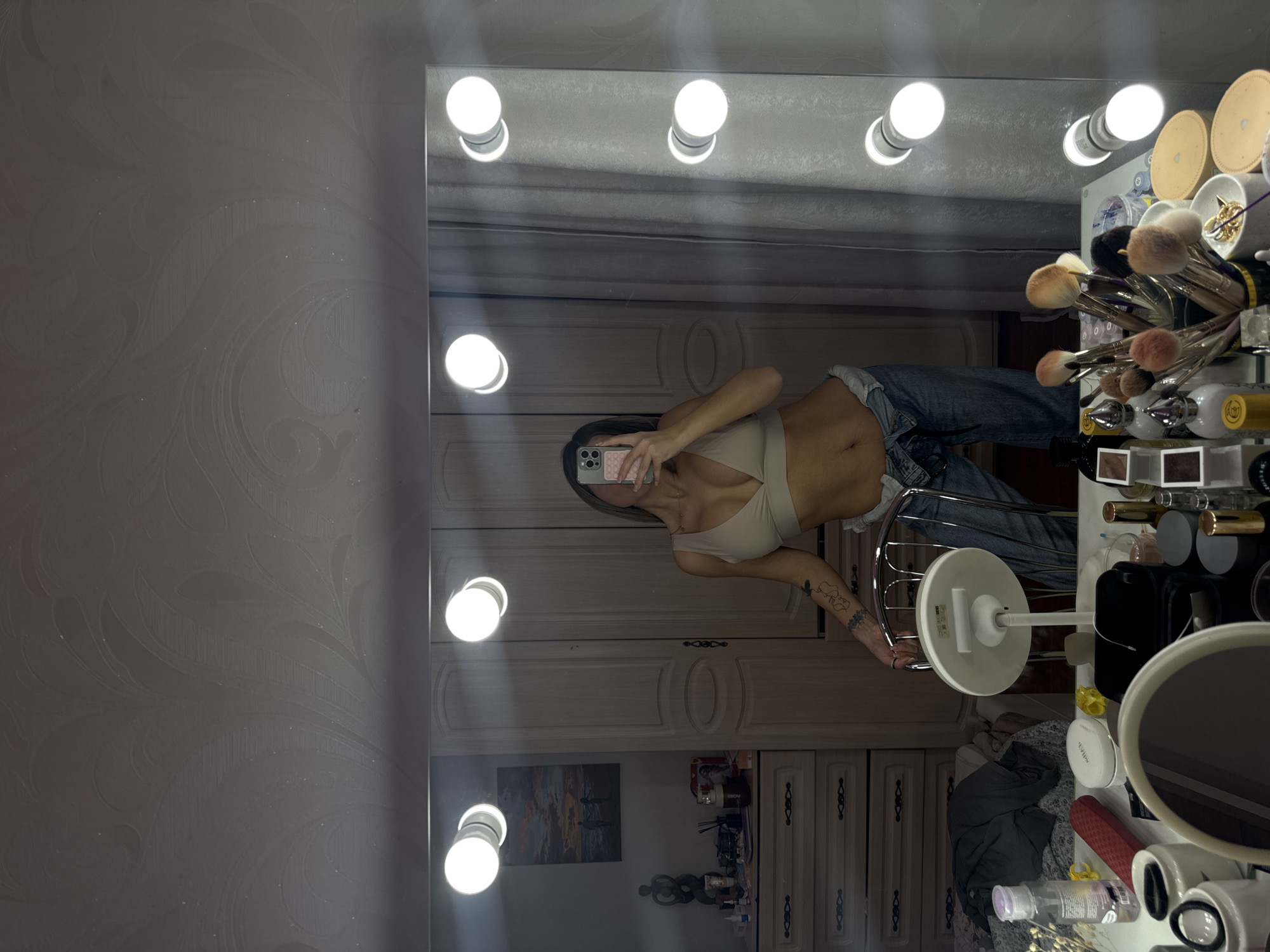 The image displays an indoor scene in a room located in Chaikovsky, Russia, captured as a selfie reflection in a large, rectangular vanity mirror. A female subject stands in the center, partially obscuring her face with a smartphone held in her right hand as she takes the photograph. She wears a beige bralette or crop top, exposing her midriff, and loose-fitting blue jeans. Her left arm is visible, adorned with multiple dark tattoos. She leans against the chrome frame and white backrest of a chair or stool, positioned in front of the vanity table.
The vanity mirror itself is lined with multiple circular light bulbs, providing illumination; three are visible along the top edge and four along the right vertical edge.
The foreground, constituting the surface of the vanity table, is cluttered with numerous cosmetic products. These include various bottles, jars, compacts, perfume bottles, and makeup brushes housed in multiple containers. A small, round magnifying mirror on a white stand is centrally located on the table, alongside a red elongated case or container and a white oval container.
The background, reflected in the mirror, reveals elements of a room. To the left, a light-colored wooden dresser with multiple drawers and decorative handles is visible. On its surface rests a framed artwork depicting two figures on a beach at sunset, a dark sculpted figurine, and additional cosmetic items. Behind the subject and to the right, a light-colored wooden wardrobe with two paneled doors is present. Further right, light-colored curtains are drawn. The walls and ceiling exhibit a light-colored wallpaper with a subtle, swirling, or floral pattern.