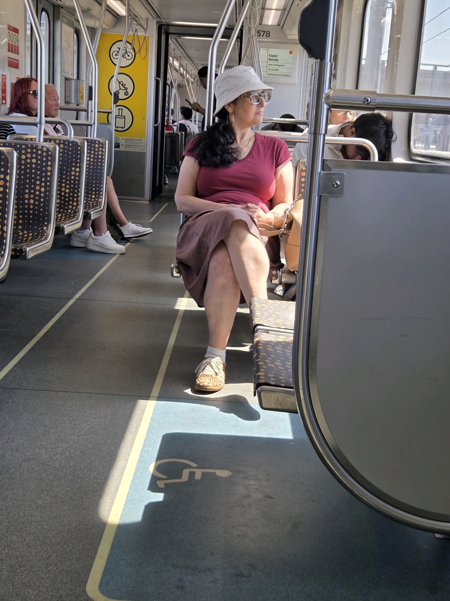 An interior shot of a Metro E Line (Expo) train car shows passengers seated and standing. The train is likely traveling eastbound on the Expo Line, as indicated by the digital display showing "Expo/Bundy" as the destination. The focus is on a woman in the foreground wearing a burgundy t-shirt, a patterned skirt, a white bucket hat, and glasses. She is seated with her legs crossed, and a tan purse is on her lap. Sunlight streams through the windows, casting shadows on the floor. Other passengers are visible in the background, including a woman with red hair and a man with gray hair seated behind her. A stroller icon is visible on a yellow sign inside the train. The train car's interior features gray seats with a dark, speckled pattern, metal poles for support, and yellow and blue lines on the floor. A wheelchair accessible space is marked on the floor with a stencil. The overall scene depicts a typical commute on Los Angeles public transportation.