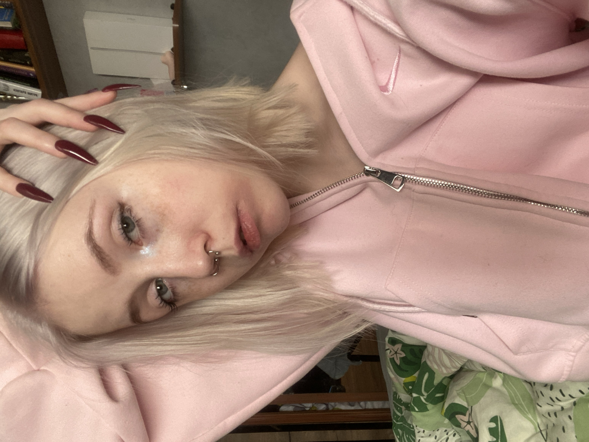 A close-up portrait features a young person, likely female, with pale skin, light blonde hair, and green-grey eyes, lying down indoors. She has visible freckles across her face and a silver horseshoe-shaped septum piercing. Her lips are naturally pink and slightly parted.
She is wearing a light pink zip-up hoodie, which has a subtle, embroidered Nike swoosh logo on the right side of the chest (from the viewer's perspective). Her left hand is raised near her head, prominently displaying her long, pointed (stiletto or almond shape) fingernails, which are painted a dark red or burgundy color.
The background suggests an indoor setting, likely a personal room such as a bedroom, given the presence of a patterned green, white, and black duvet or blanket in the lower right corner. A stack of books is visible on a shelf in the upper left, with some Cyrillic text discernable, including "КOHTAPO" (possibly part of a larger word like "Контаров" or "Контакт") and "ИГРА" (meaning "game"). This detail aligns with the provided location context of Izhevsk, Russia. A white rectangular object, possibly furniture, is also seen behind the books.
The person's expression is neutral and relaxed, with her gaze directed towards the camera. The lighting is soft and even, indicating indoor conditions without direct sunlight. The image appears to be a selfie or a closely framed portrait.