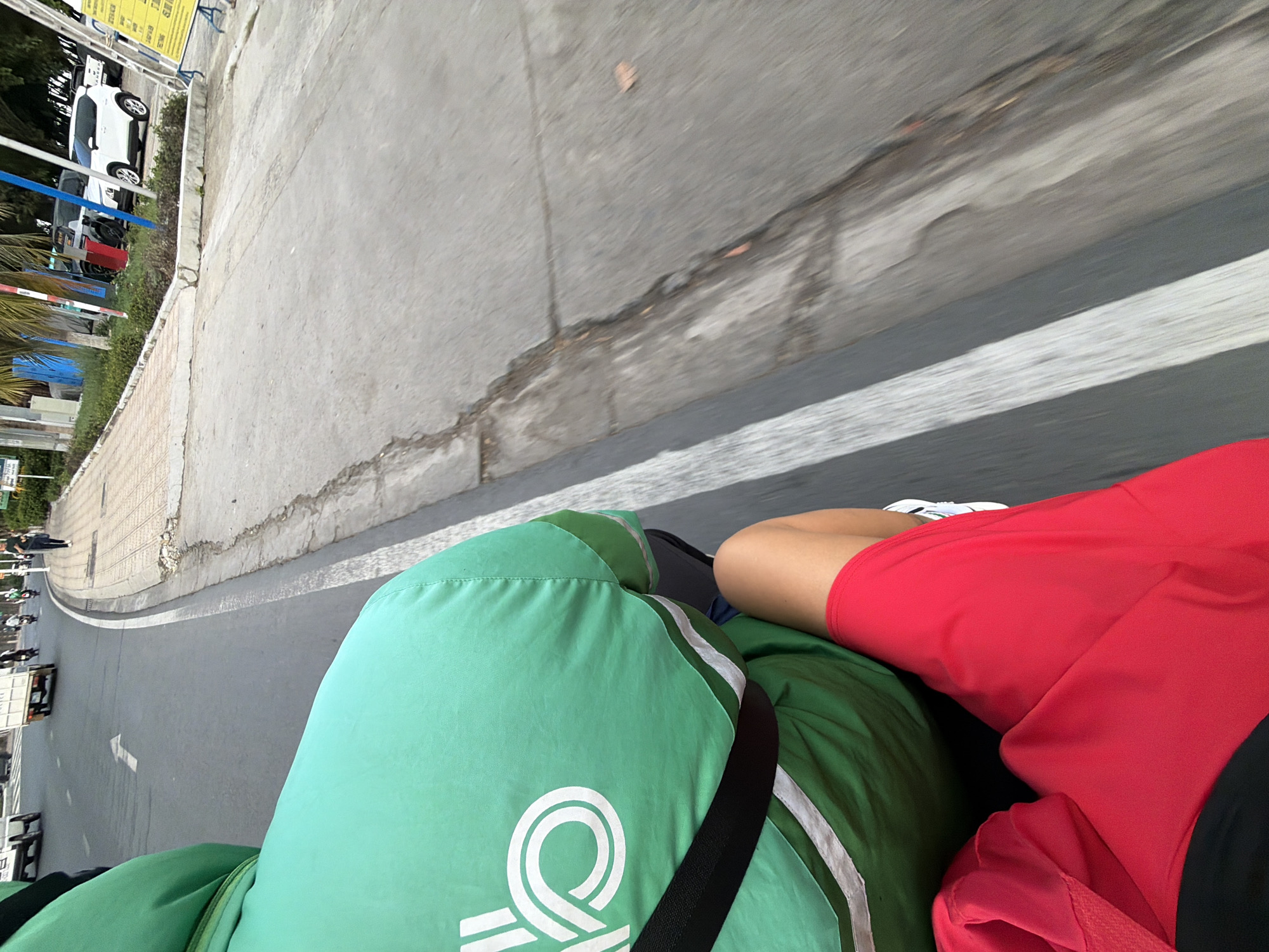 A first-person perspective shot taken from the back of a motorcycle in Nha Trang, Vietnam. The rider is wearing a bright green hooded jacket with white stripes and a logo, and red shorts. The motorcycle is moving along a paved road that appears to be in moderate condition, with some cracks and patches visible. To the right of the road is a sidewalk made of light-colored tiles, bordered by a low concrete wall. Beyond the sidewalk, there are palm trees, some parked cars, and buildings. The overall scene suggests a daytime urban environment in Vietnam.