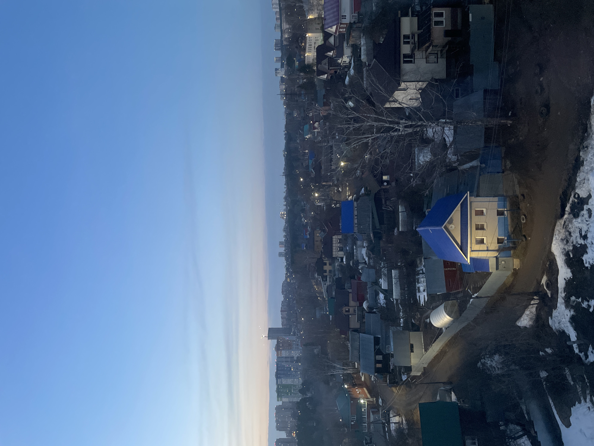 The image shows an elevated view of a residential area in Izhevsk, Russia. The left side of the frame is dominated by a pale blue sky. Moving to the right, the scene shifts to an urban landscape of varying elevations, with an abundance of houses featuring different roof colors and styles. Some buildings appear to be low-rise apartments, especially in the background. A prominent bare tree stands out amidst the buildings. Snow patches are visible near a road on the right side, suggesting winter or early spring. Streetlights illuminate the scene, indicating either early morning or late evening. The foreground houses appear to be more individualized dwellings.