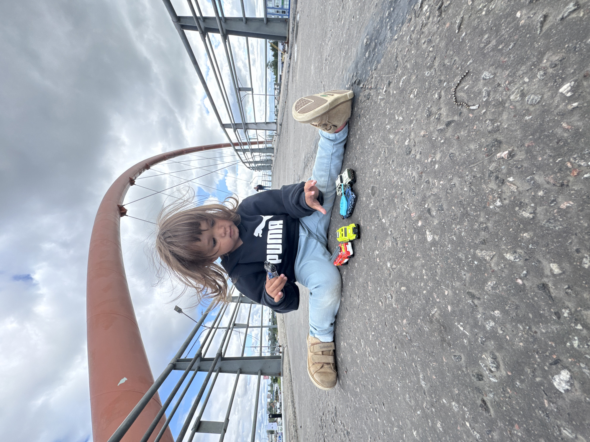 A young child with brown hair, wearing a black Puma hoodie and light blue jeans, sits on the asphalt surface of a pedestrian bridge with their legs spread in a straddle split. The child is playing with three toy cars: a red and white fire truck, a yellow car, and a blue car. The child's left foot is extended outwards, and their right foot is bent. The bridge has a metal railing on either side, and a large, orange, arched structure is visible in the background. The sky is cloudy. The scene appears to be in Saint Petersburg, Russia, given the architectural style of the bridge and the surrounding urban environment.