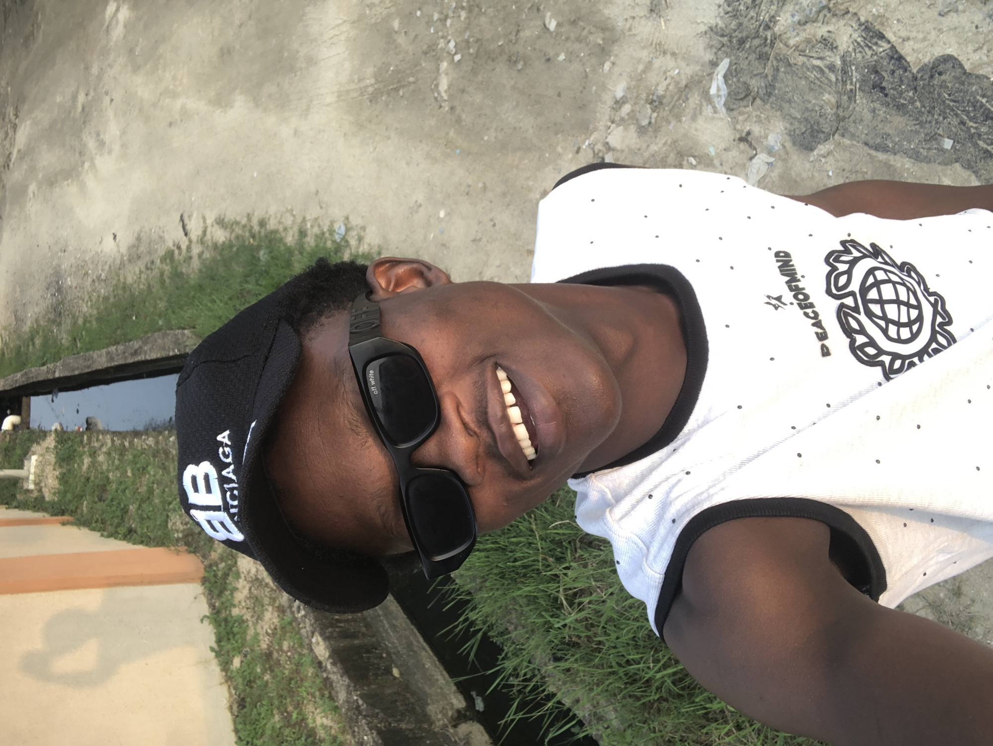 The image shows a close-up, selfie-style shot of a young Black male outdoors. He is wearing a black baseball cap with white lettering that appears to say "BB NCIAGA" and black sunglasses. His white sleeveless shirt has a black trim and a small pattern of black dots. A graphic design featuring the words "PEACEOFMINND" and a globe-like image is visible on his shirt. He is smiling, showing his teeth.
The background suggests an outdoor setting. To the left, there's a textured wall, possibly painted orange and cream, with some green vegetation and a concrete ditch with water. To the right, the ground appears to be unpaved, with dirt and some debris. The lighting suggests it's daytime. The context provided indicates the location is in the city of Ado, Nigeria, likely an urban or peri-urban area given the presence of a built wall and drainage channel alongside unpaved ground.