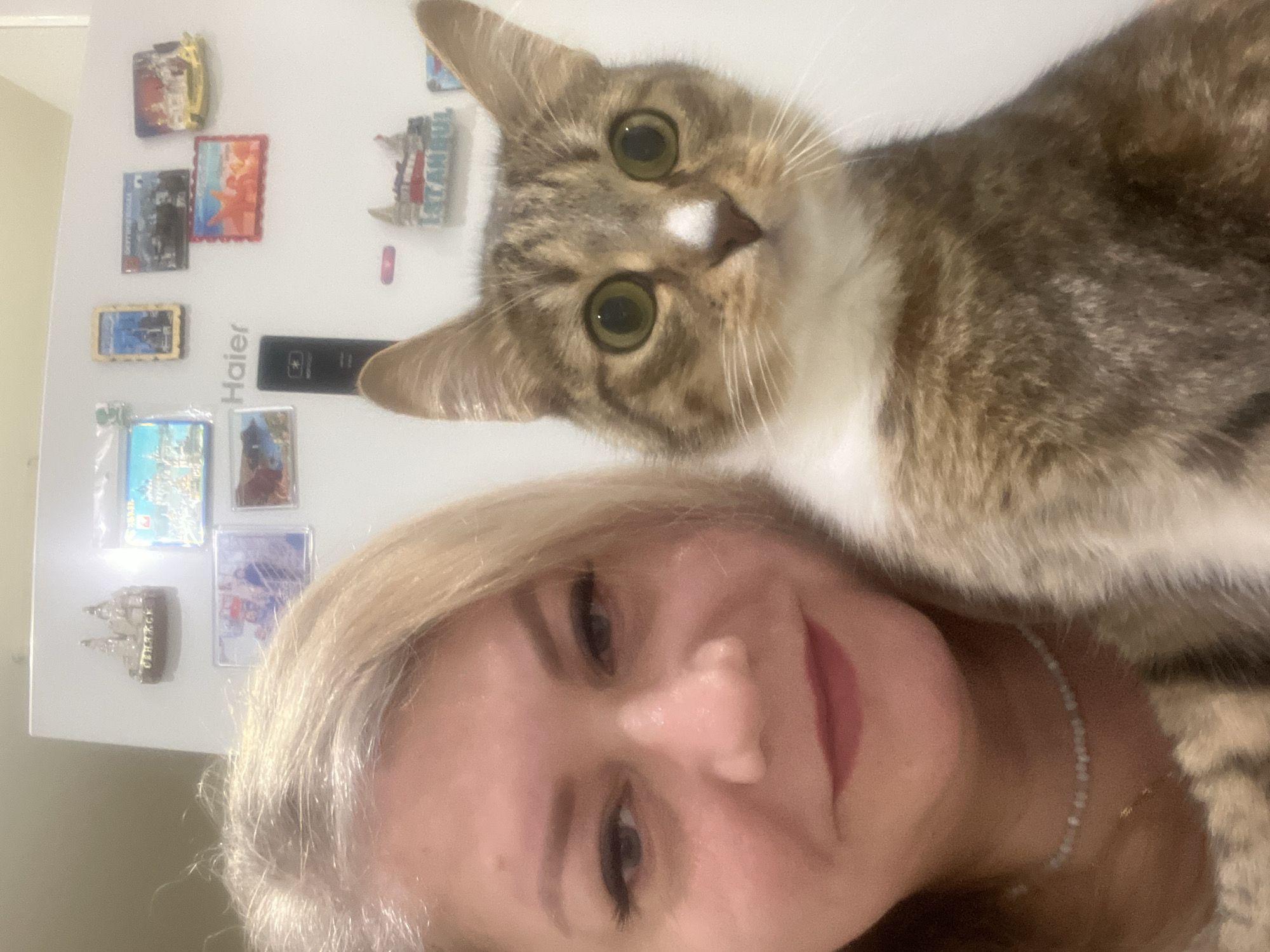 A woman with light blonde hair is in the foreground of the image, smiling at the camera. A tabby cat with a white chest and a white spot on its nose is positioned above her head, looking at the camera as well. Behind the woman and the cat, a white refrigerator is visible with several magnets attached. The magnets appear to be souvenirs from different cities and landmarks, including what might be a magnet from Istanbul. The refrigerator has a "Haier" brand logo and a control panel. The location is not identifiable as Izhevsk, Russia, based on the provided information.