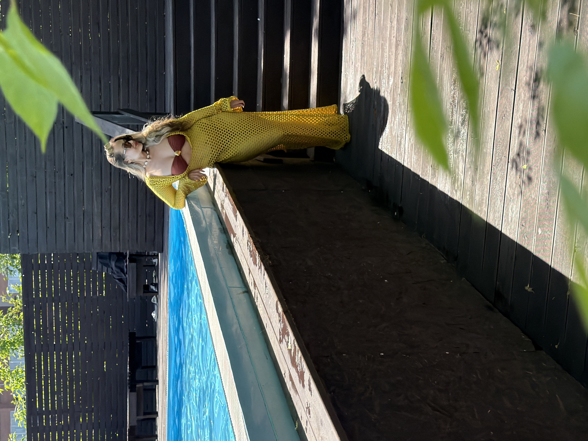 An outdoor, sunlit photograph features a woman posing by a swimming pool on a wooden deck, likely in Krasnoyarsk, Russia. The image is currently displayed on its side (rotated 90 degrees clockwise).
The central subject is a woman with long, wavy blonde hair, wearing light-colored sunglasses. She is dressed in a dark brown bikini top and a sheer, bright yellow, long-sleeved cover-up with a mesh or fishnet pattern. A necklace adorned with shell-like beads is visible around her neck. Her left arm is slightly bent, with her hand visible.
She stands on a light brown wooden deck, which shows some signs of weathering, positioned alongside a rectangular swimming pool filled with clear blue water, exhibiting ripples on its surface. In the background, behind the woman, there is a dark wooden fence or wall composed of vertical slats.
The scene is illuminated by bright sunlight, casting long, distinct shadows across the wooden deck, including a prominent shadow of the woman. Out-of-focus green leaves are visible in the foreground, subtly framing the top left and right portions of the image. The overall impression is one of a relaxed, sunny outdoor setting. There is no visible text in the image.
