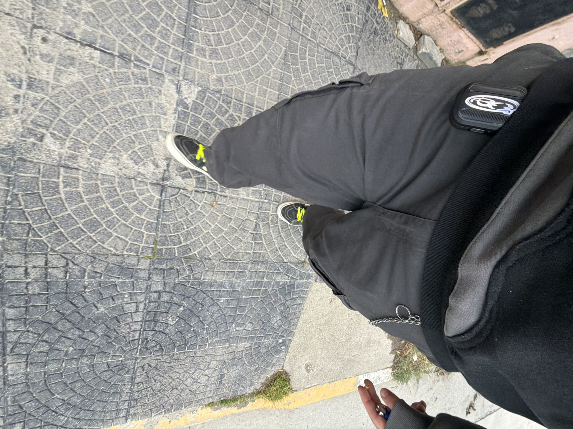 A first-person, downward-angled shot shows a person standing on a sidewalk composed of grey, circular-patterned paving stones. The person is wearing dark grey cargo pants with a dark grey sweatshirt and black and white sneakers with neon yellow accents. An Airpods case with a graphic is clipped onto the right pocket of the pants. A chain is visible, hanging from the pants' left pocket. The lower portion of the person's left hand is visible in the bottom right corner. The sidewalk transitions to a concrete curb edged with yellow. Patches of grass grow along the curb. In the upper right corner, there is a grey concrete structure with inscriptions. Based on the paving stones and the structure, the location is likely the city of Ushuaia, Argentina.