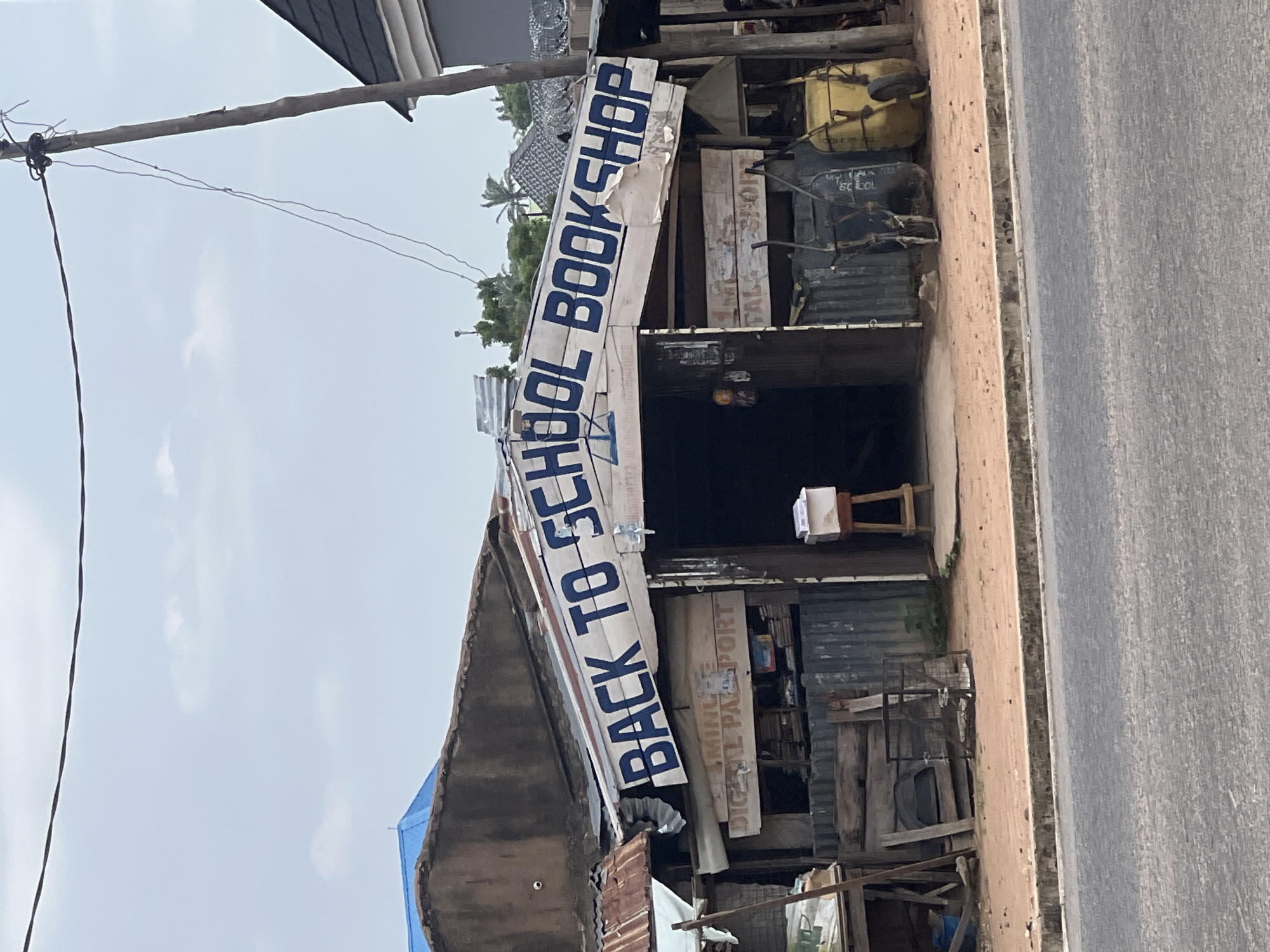 The image depicts a roadside "Back to School Bookshop" in Jalingo, Nigeria. The shop has a corrugated metal roof and an open storefront. A white banner with blue lettering that reads "BACK TO SCHOOL BOOKSHOP" is prominently displayed across the front of the roof. Below the banner, a sign reads "DIGITAL PASSPORT". Various items, including books, are visible inside and around the shop. Two wheelbarrows, one yellow and one dark-colored, are parked in front of the shop. A wooden stool with a white box on top is placed near the entrance. The shop is situated on a dirt road, with a paved road in the foreground. The environment appears to be a commercial area with other buildings visible in the background. There are no people clearly visible in the image.