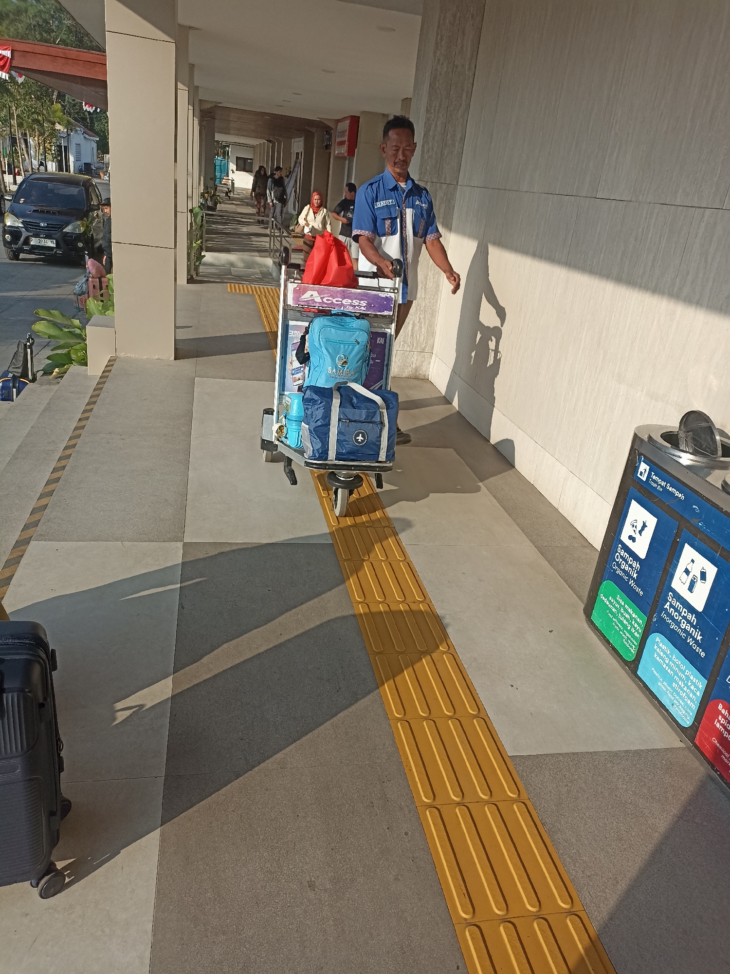 The image depicts a man wearing a blue uniform with "Access" and "KAI" logos, pushing a luggage cart laden with bags. He appears to be an employee at the Mojoagung railway station in Indonesia, as indicated by the "KAI" (Kereta Api Indonesia) logo, which signifies the Indonesian state-owned railway company. The cart carries a bright red bag, a blue backpack, and a blue duffel bag. The man is walking along a walkway with tactile paving, characteristic of a public transportation hub. To the right, a multi-compartment trash bin is visible, labeled in Indonesian for organic and inorganic waste. In the background, a black vehicle, possibly a car, is parked. The scene is illuminated by sunlight, casting long shadows on the ground, suggesting it's either morning or late afternoon. The environment appears to be an outdoor, covered walkway at a train station.
