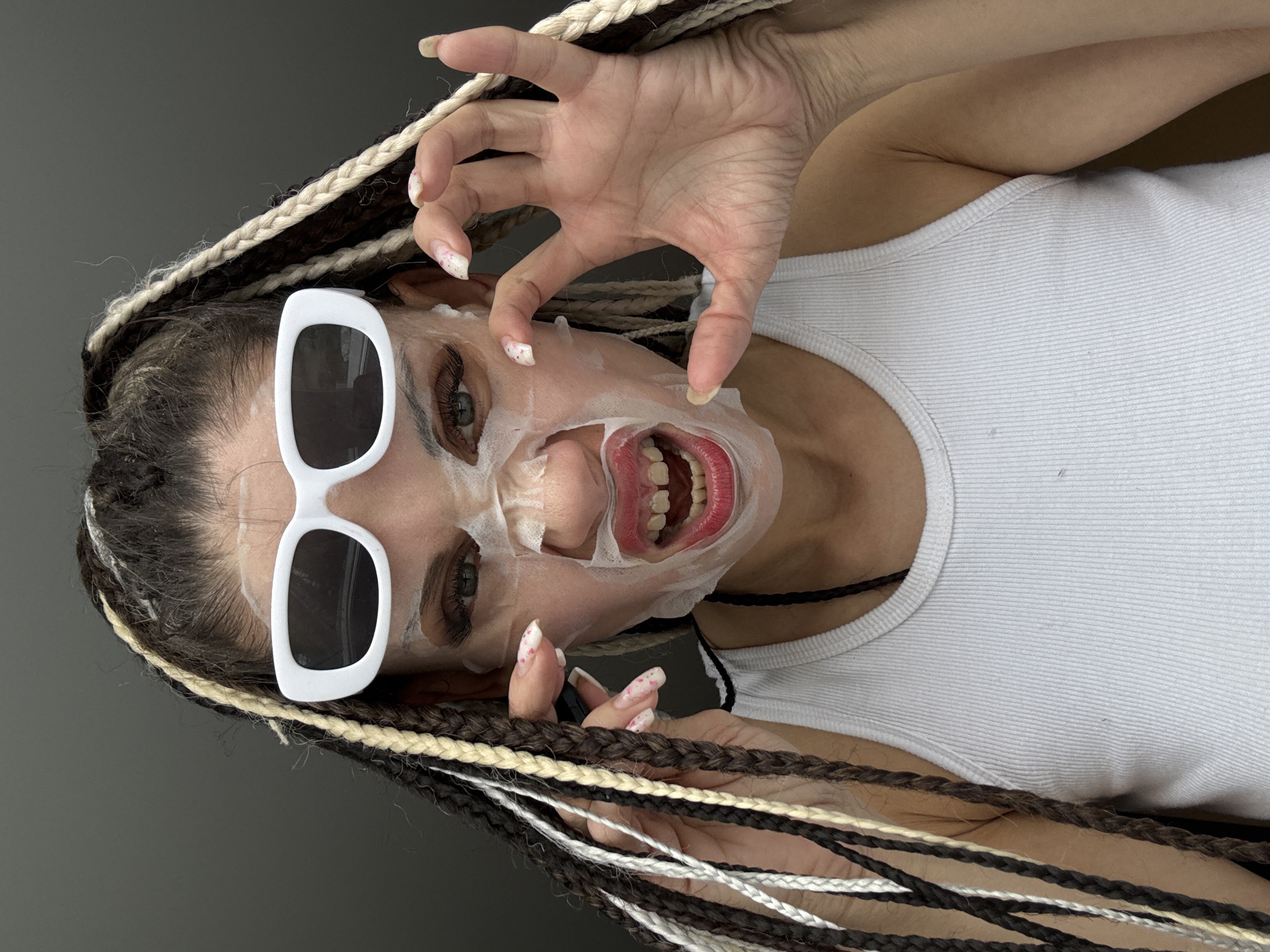 A person is lying on their back with a white sheet mask partially removed from their face, revealing their mouth with red lipstick. The person is wearing white-framed sunglasses and a white tank top. They have braided hair with brown, black, and white strands. One hand is raised in a claw-like gesture near their face. The background is a solid gray color. The location is not evident from the image.