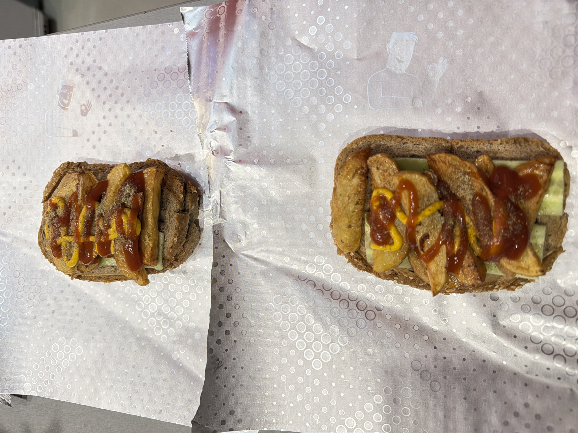 Two sandwiches are on white patterned paper. The sandwiches appear to have french fries, ketchup, and mustard. The location is likely a restaurant or food establishment, potentially in Varna, Bulgaria. There are no people visible in the image.